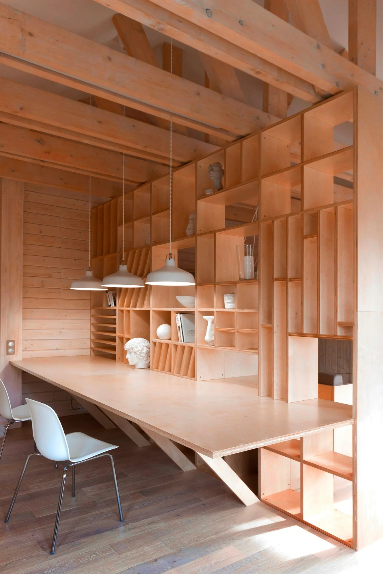 A minimalist home office features a long custom plywood desk integrated with gridded wooden shelves beneath exposed timber ceiling beams.