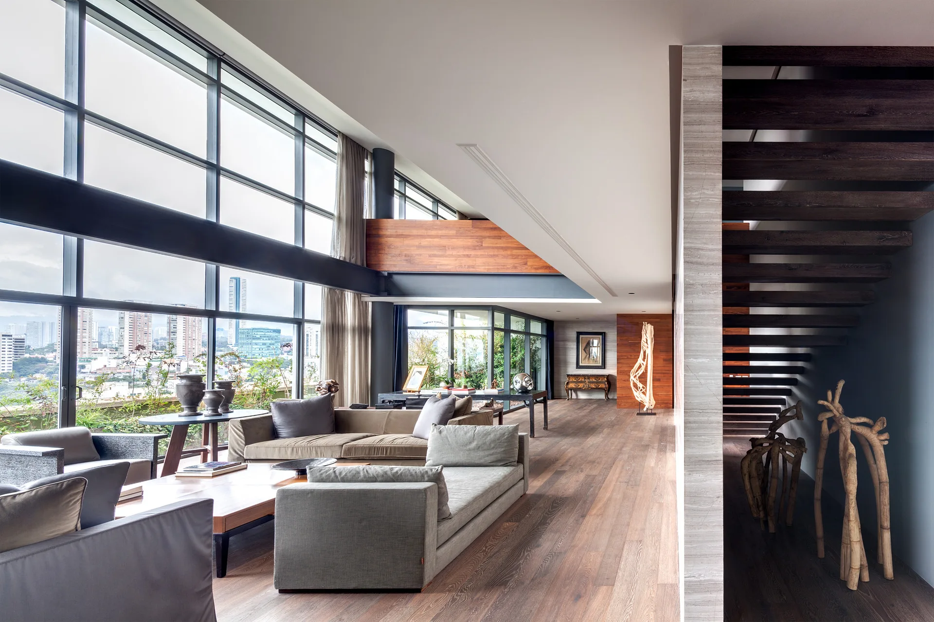 A contemporary living room with low-profile grey sofas, wood flooring, an open wood staircase, and large windows overlooking a city skyline.