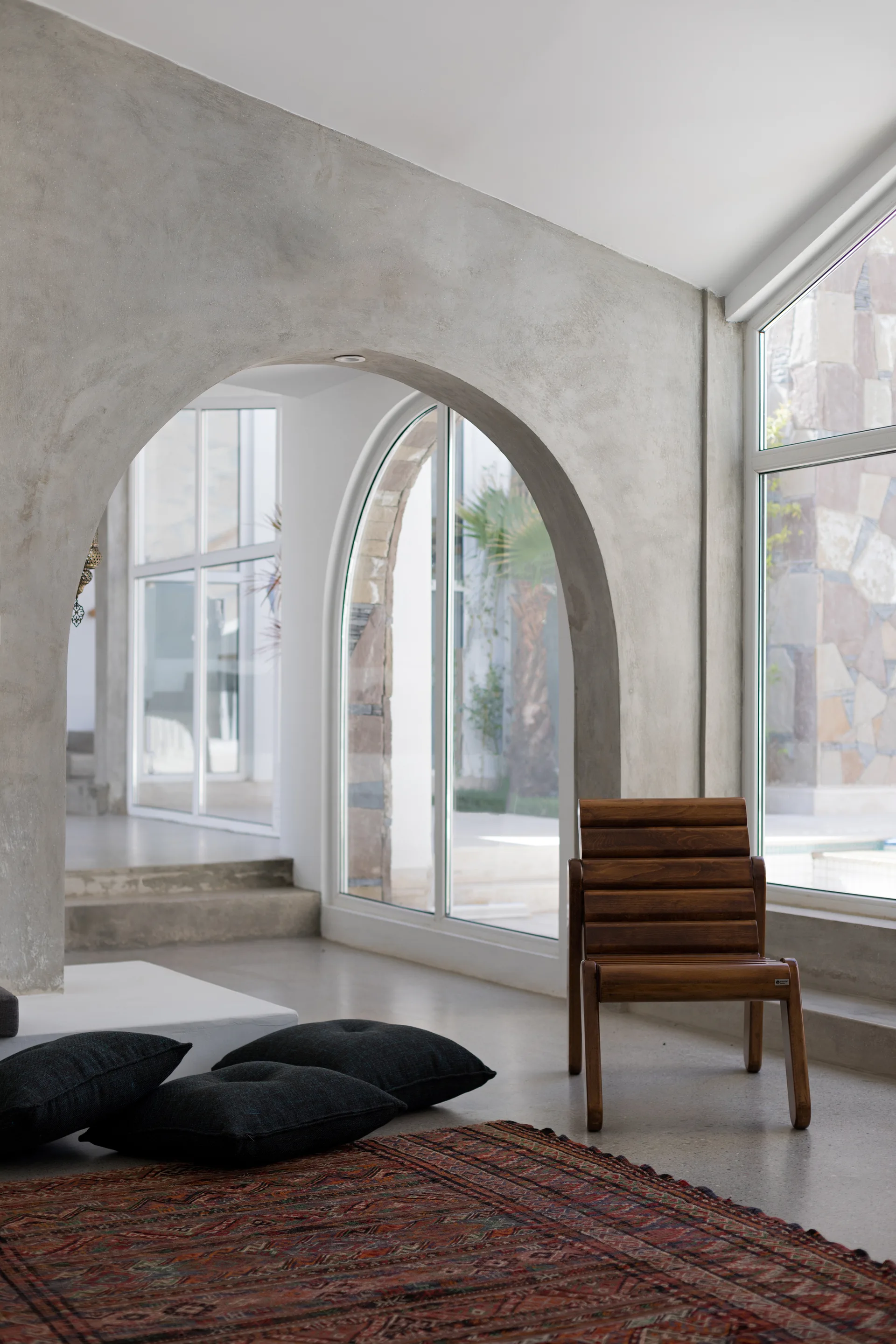 A wooden armchair and dark floor cushions sit near a textured concrete archway and arched glass windows in a minimalist room.