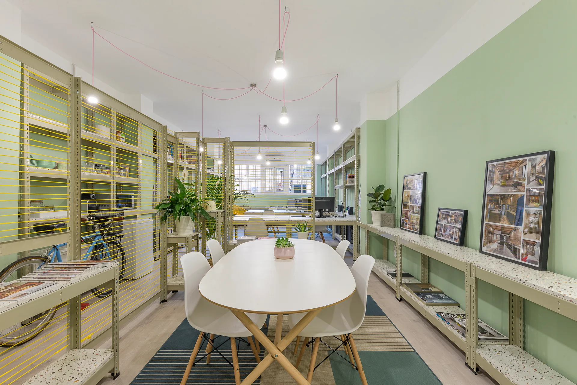 An open-plan workspace featuring an oval meeting table, white chairs, sage green walls, and industrial metal shelving units with yellow cord accents.