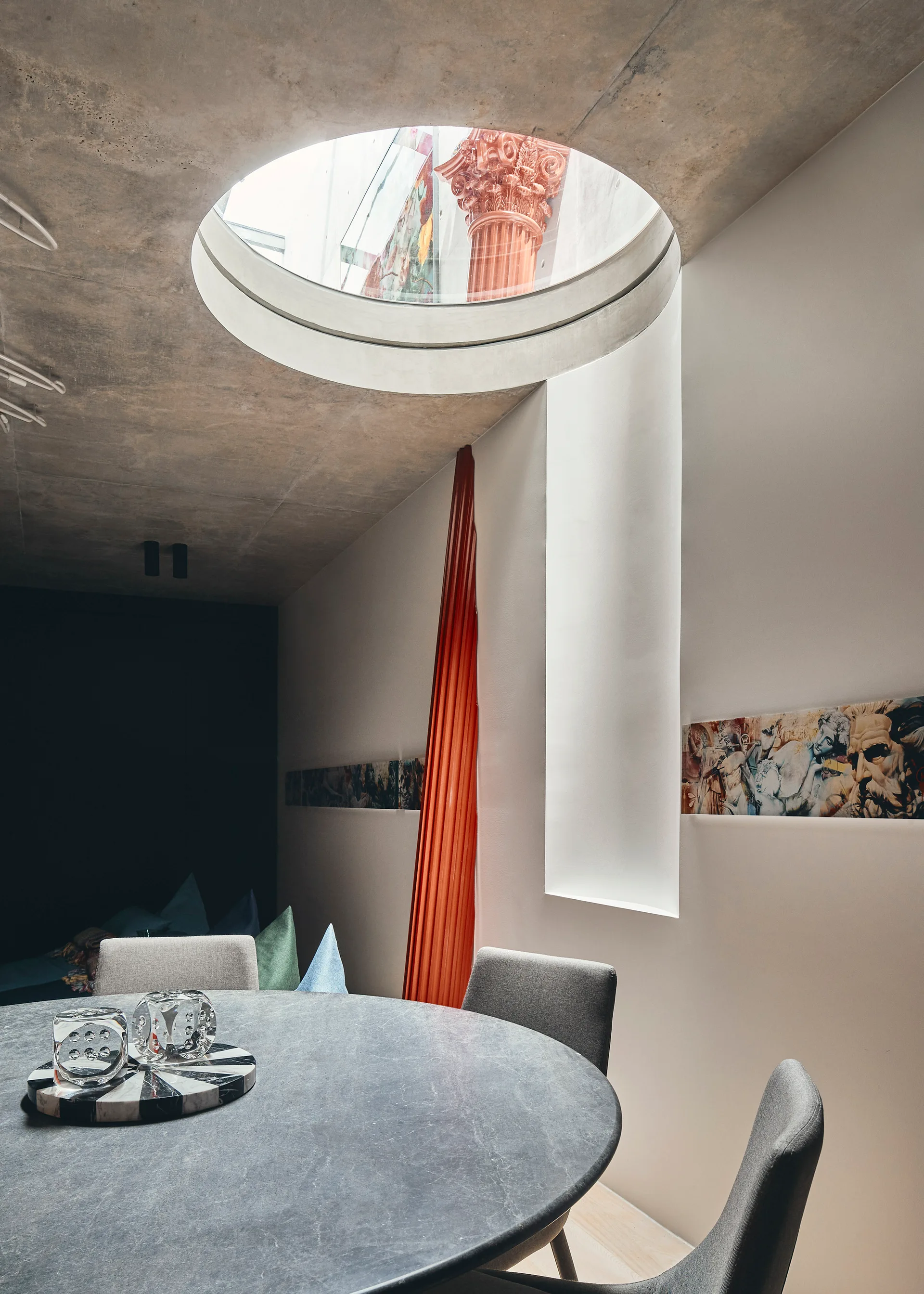 A modern dining room features a round stone table and grey chairs under a raw concrete ceiling with a large circular skylight.
