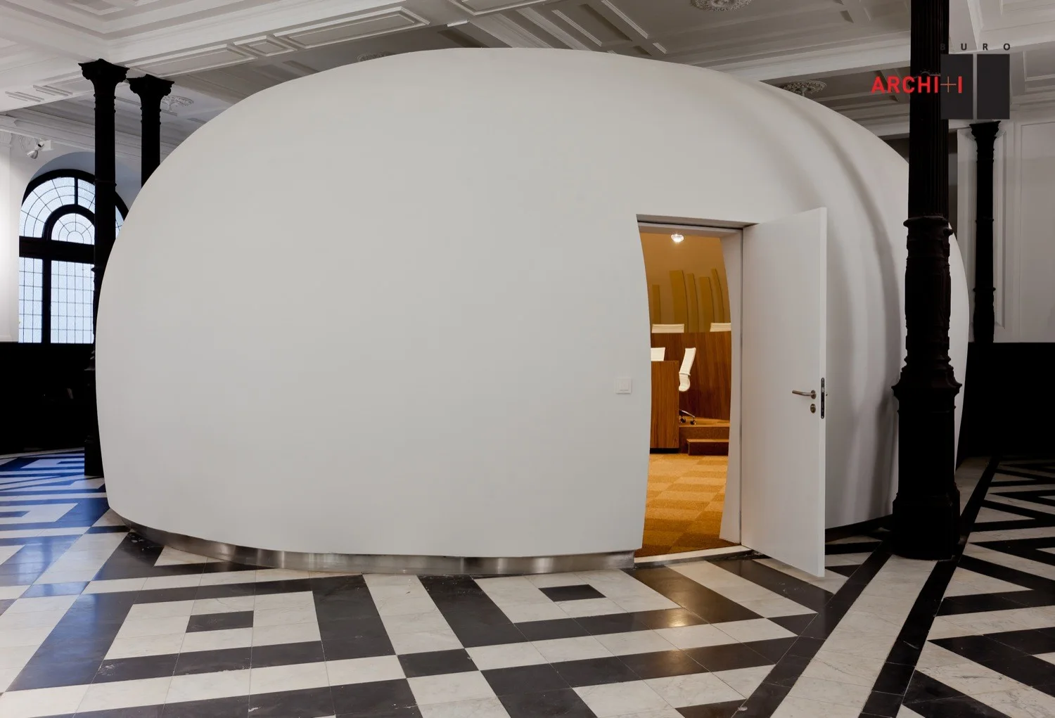 A large white dome-shaped office pod with an open door stands on a black and white checkered tile floor inside a historic space.
