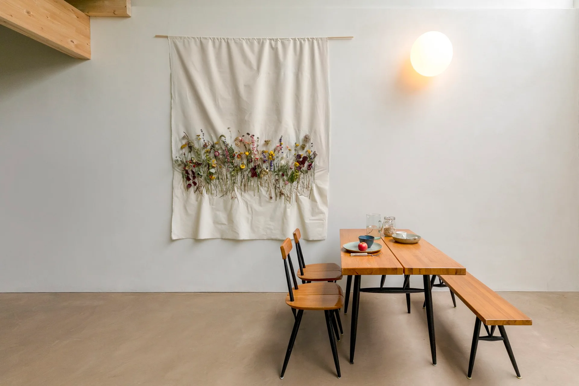 A wooden dining table with chairs and a bench sits against a white wall decorated with a fabric tapestry of dried flowers and a round glowing sconce.