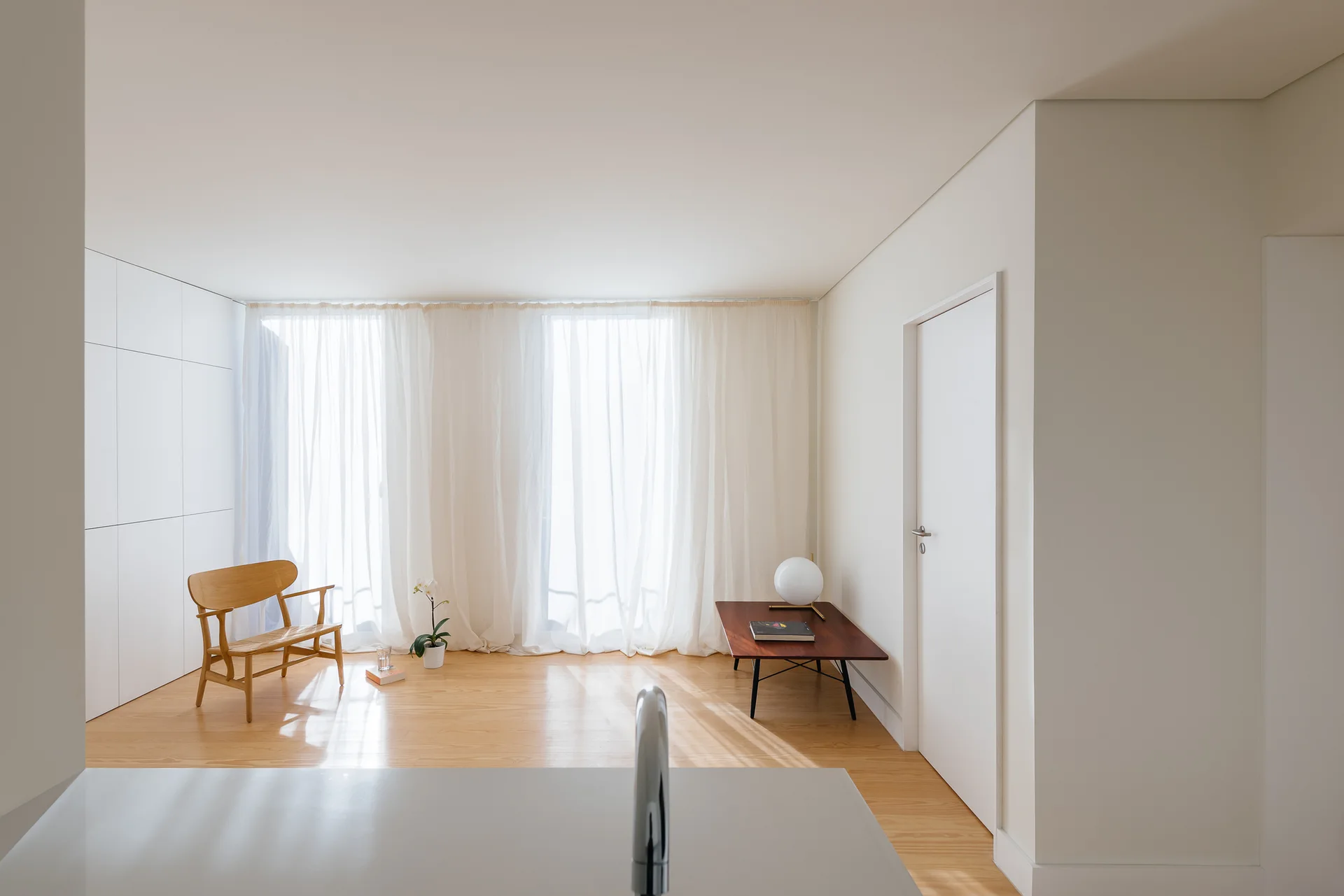 A bright minimalist room features a wooden armchair, a low wood side table with a spherical lamp, and sheer white floor-to-ceiling curtains.