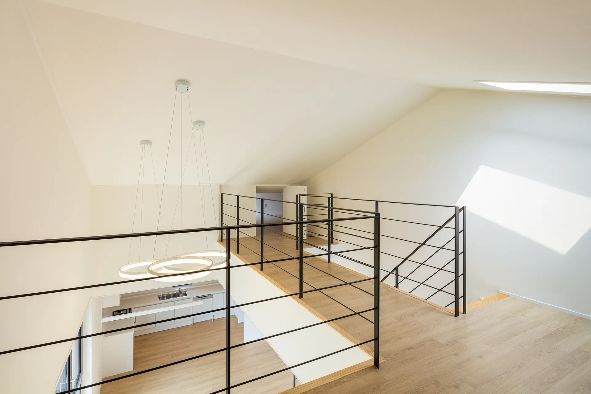 High-angle view of a modern mezzanine landing with light wood floors, a black metal railing, and a sloped ceiling overlooking a kitchen below.