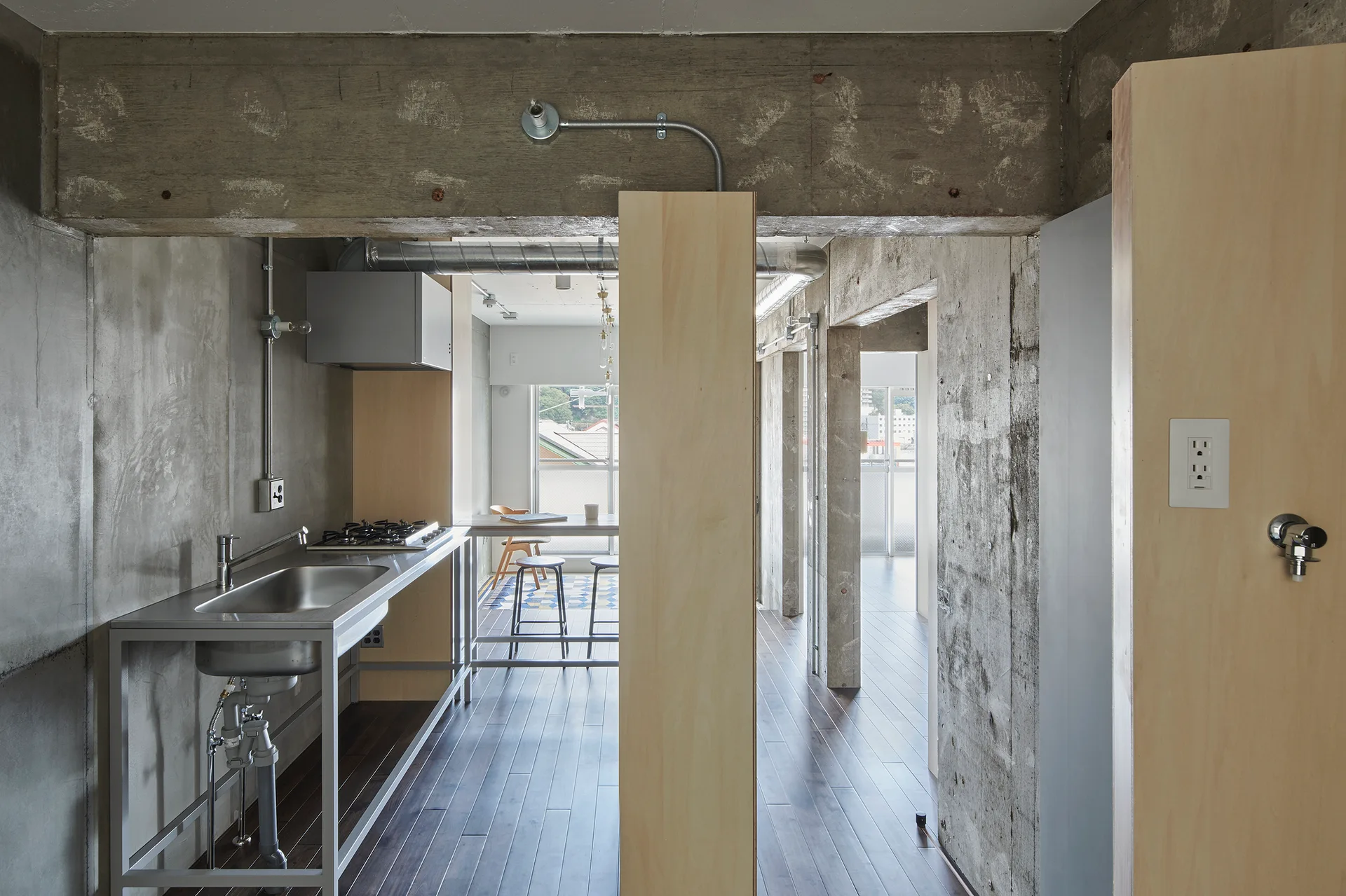 An industrial kitchen features exposed concrete walls, a stainless steel sink unit, and tall light plywood partitions on dark wood flooring.