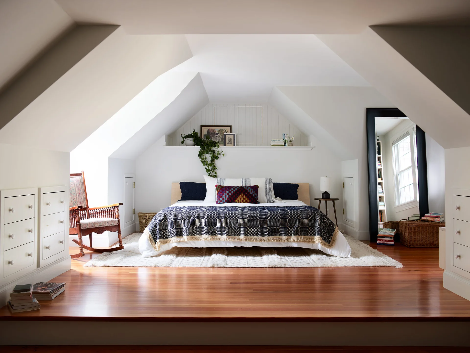 An attic bedroom featuring a bed centered in an alcove with slanted ceilings, white walls, symmetrical built-in dressers, and a large floor mirror.
