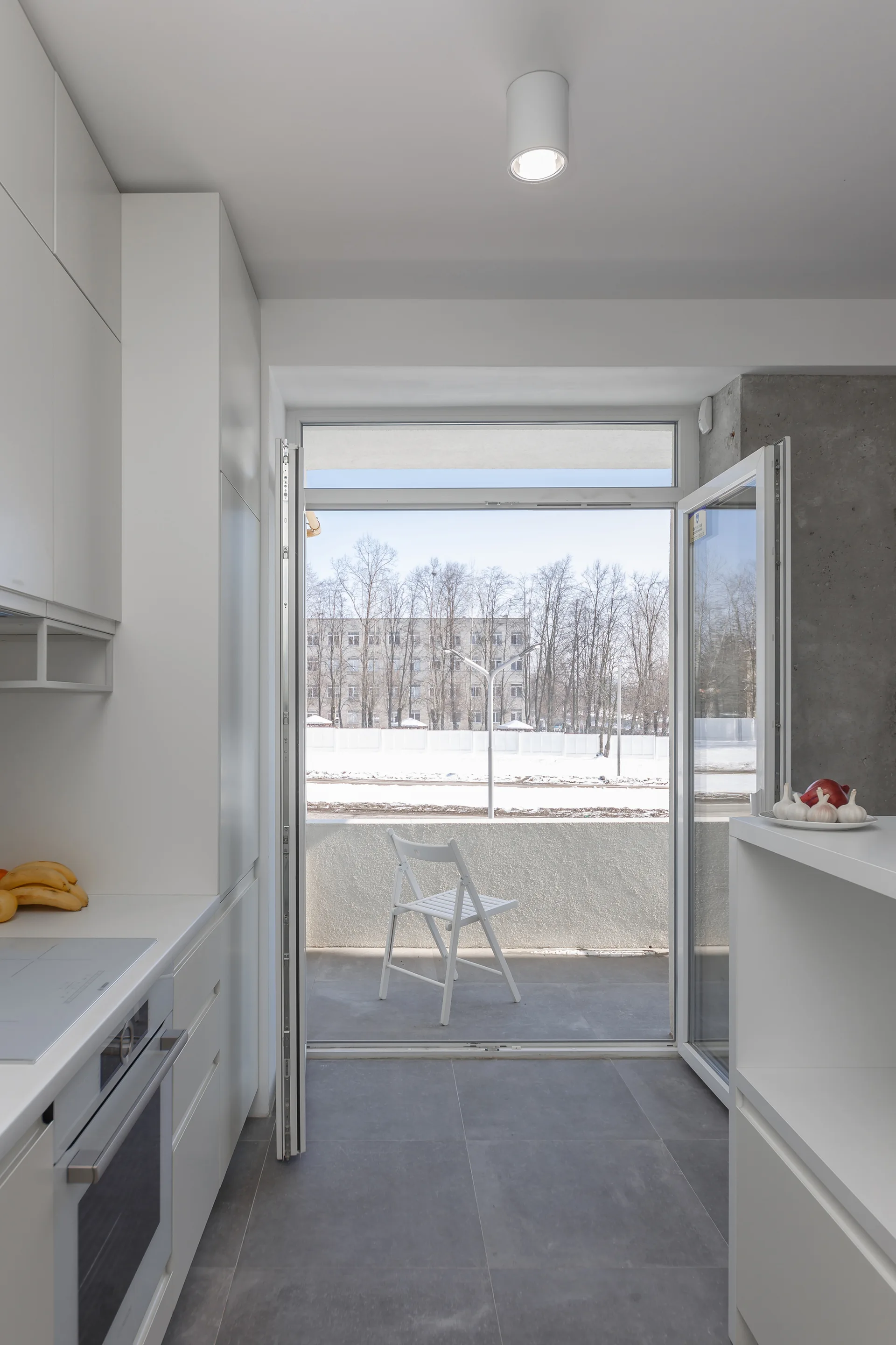 A minimalist white kitchen with integrated appliances features a large open glass door leading to a concrete balcony with a single folding chair.