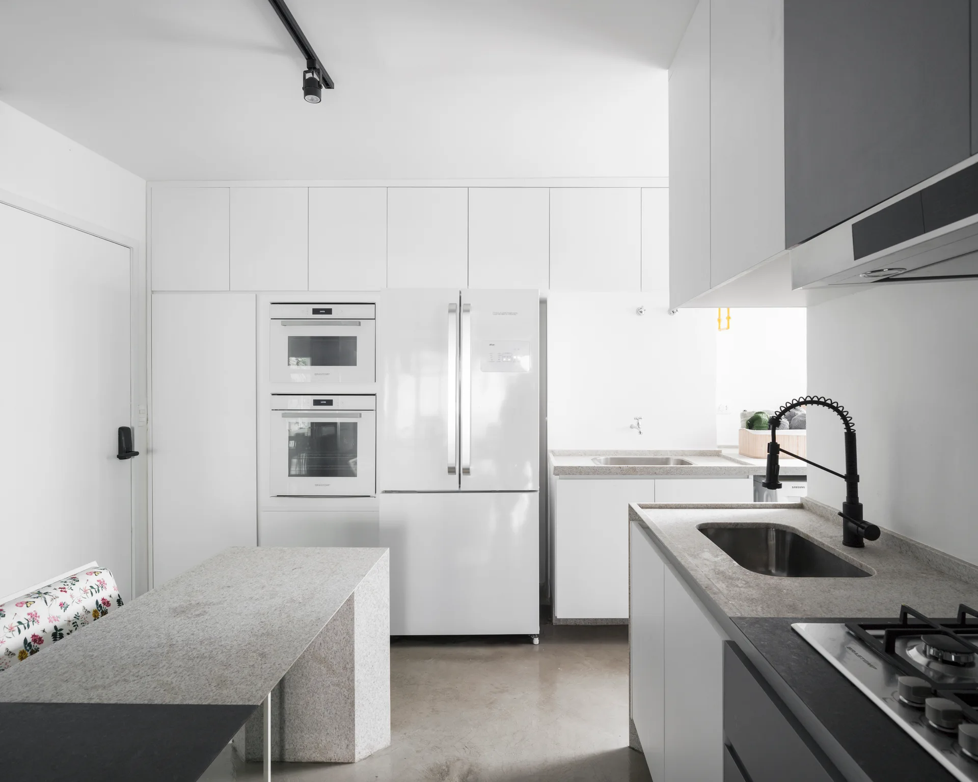 A minimalist white kitchen features flat-panel cabinets, integrated white appliances, a light stone countertop, a black gooseneck faucet, and concrete flooring.