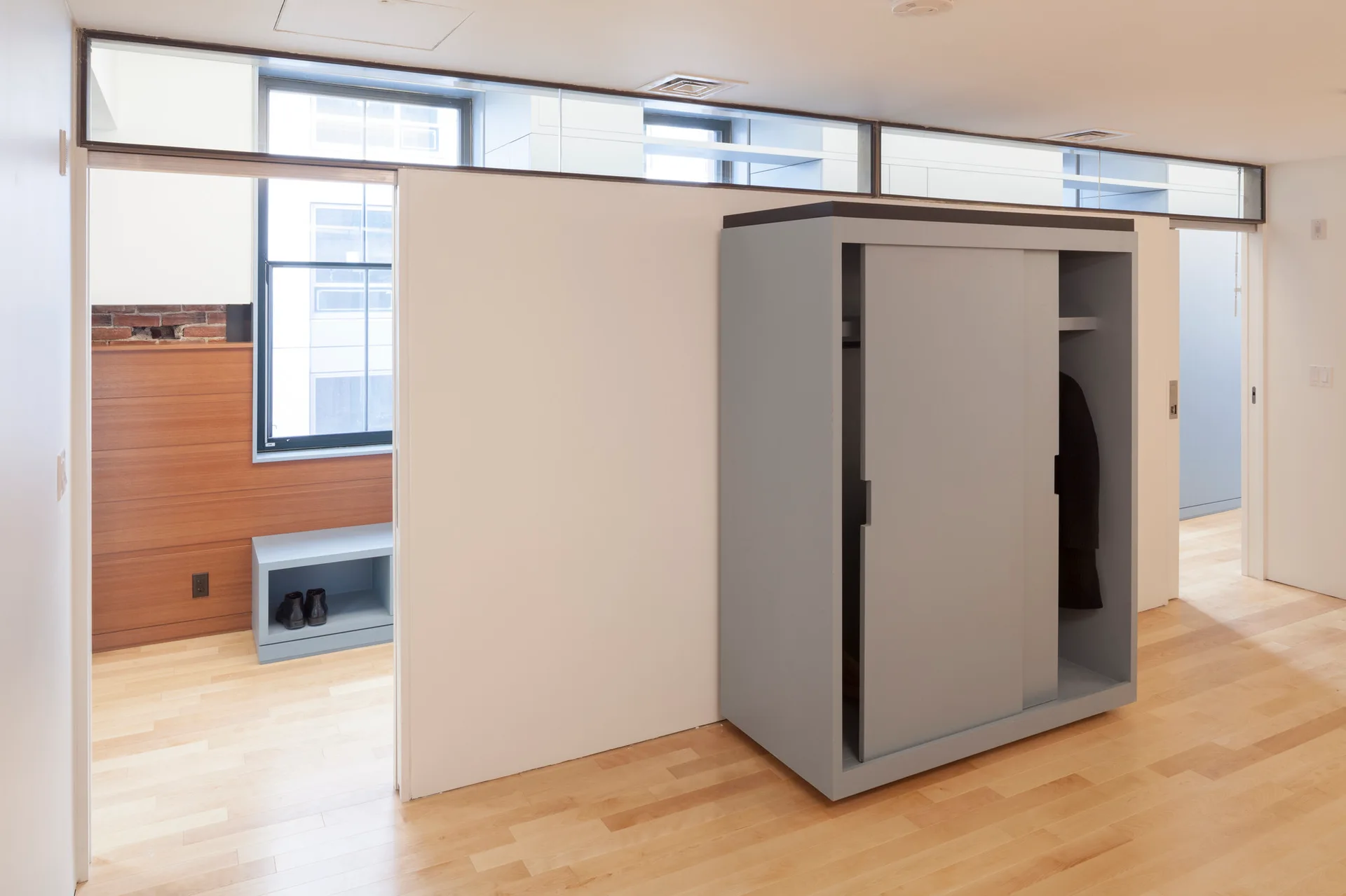 A grey freestanding wardrobe with sliding doors stands on hardwood floors next to a white partition wall with overhead transom windows.