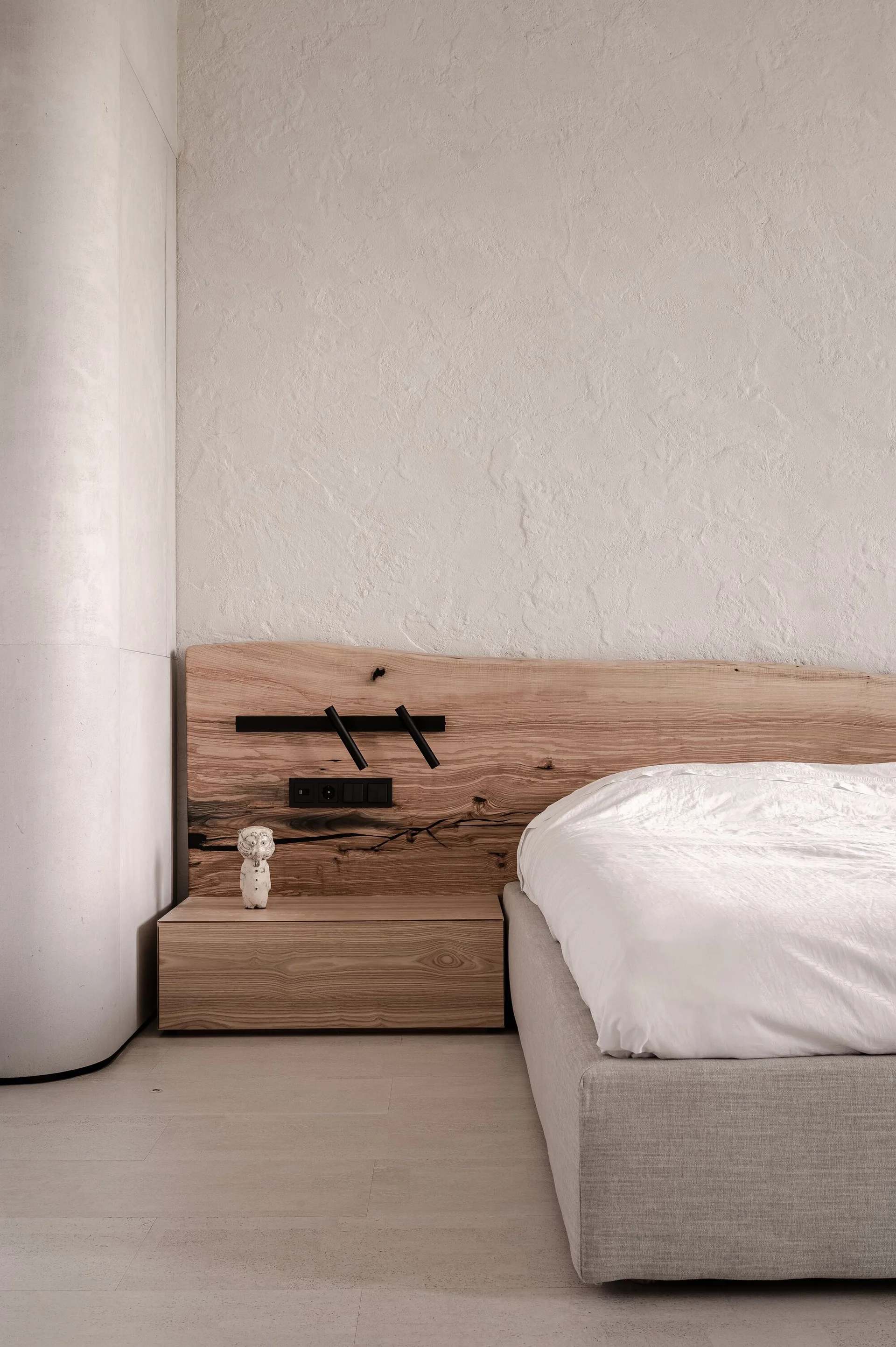 A minimalist bedroom featuring a live edge wood headboard, textured plaster walls, and a low-profile bed with a wooden floating nightstand.