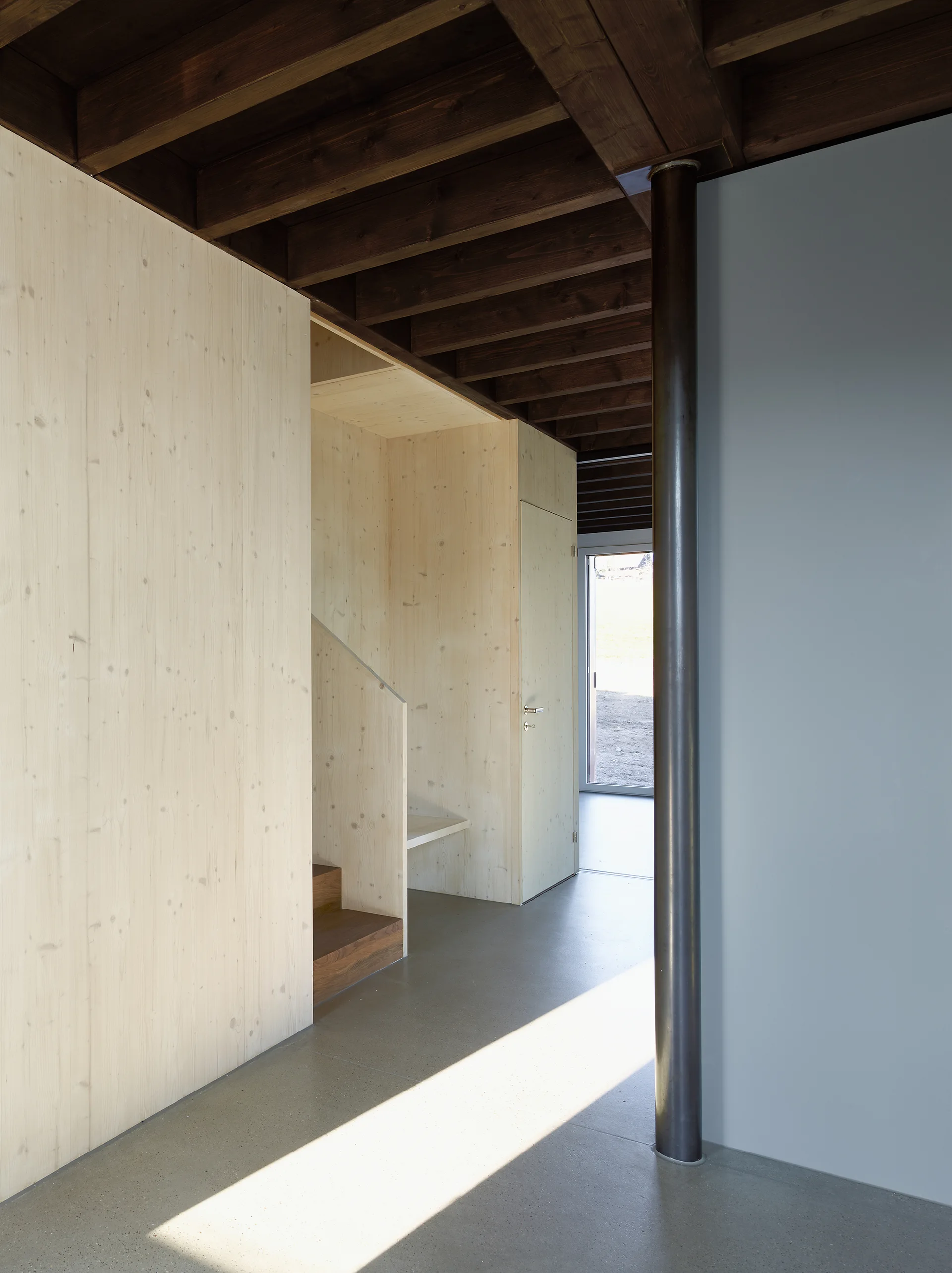 A modern entryway featuring light wood paneled walls, a nested staircase, dark exposed ceiling beams, and a smooth grey concrete floor.