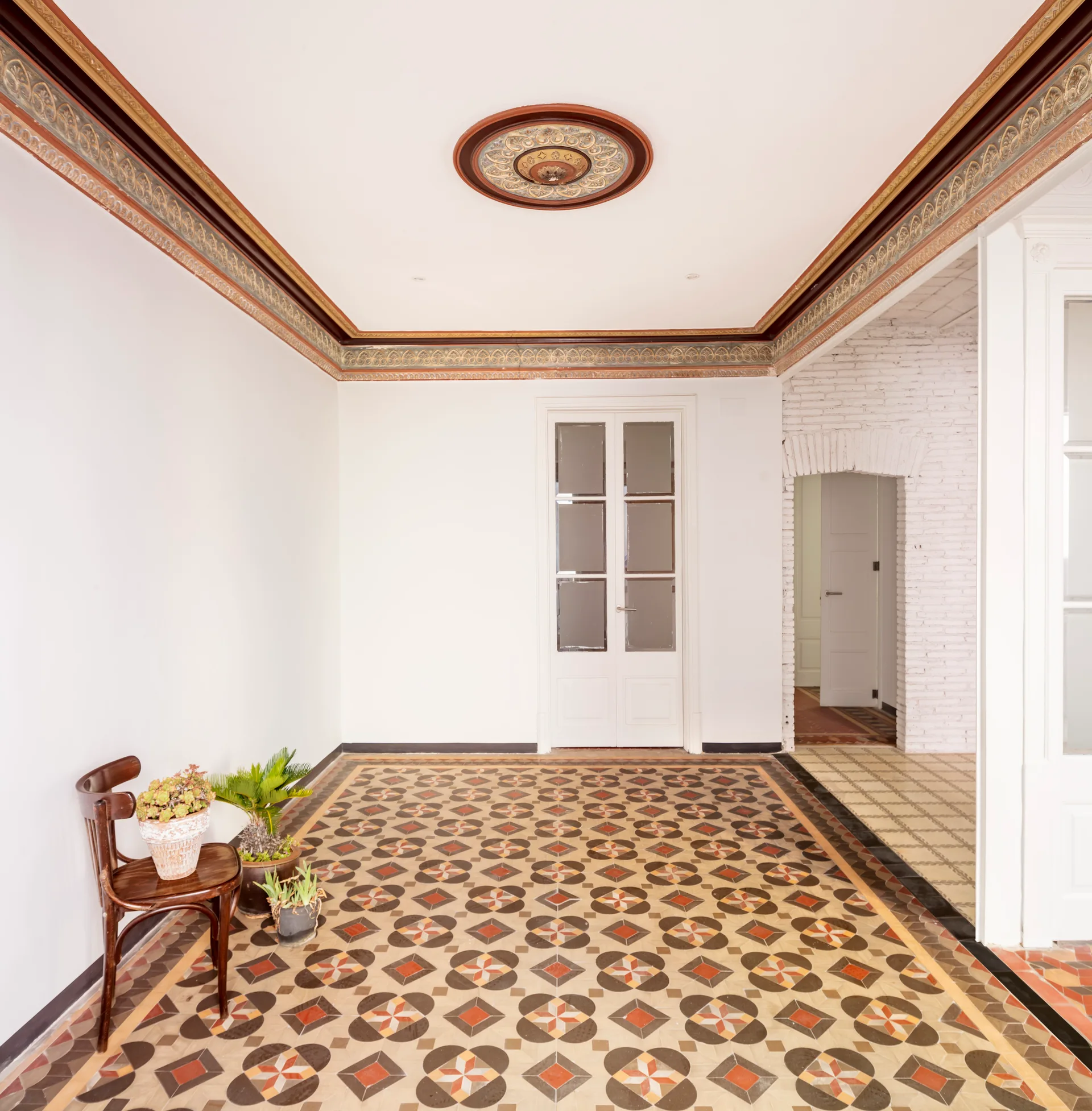 A white foyer features a highly patterned geometric tile floor, an ornate ceiling medallion, decorative crown molding, and a dark wood chair.