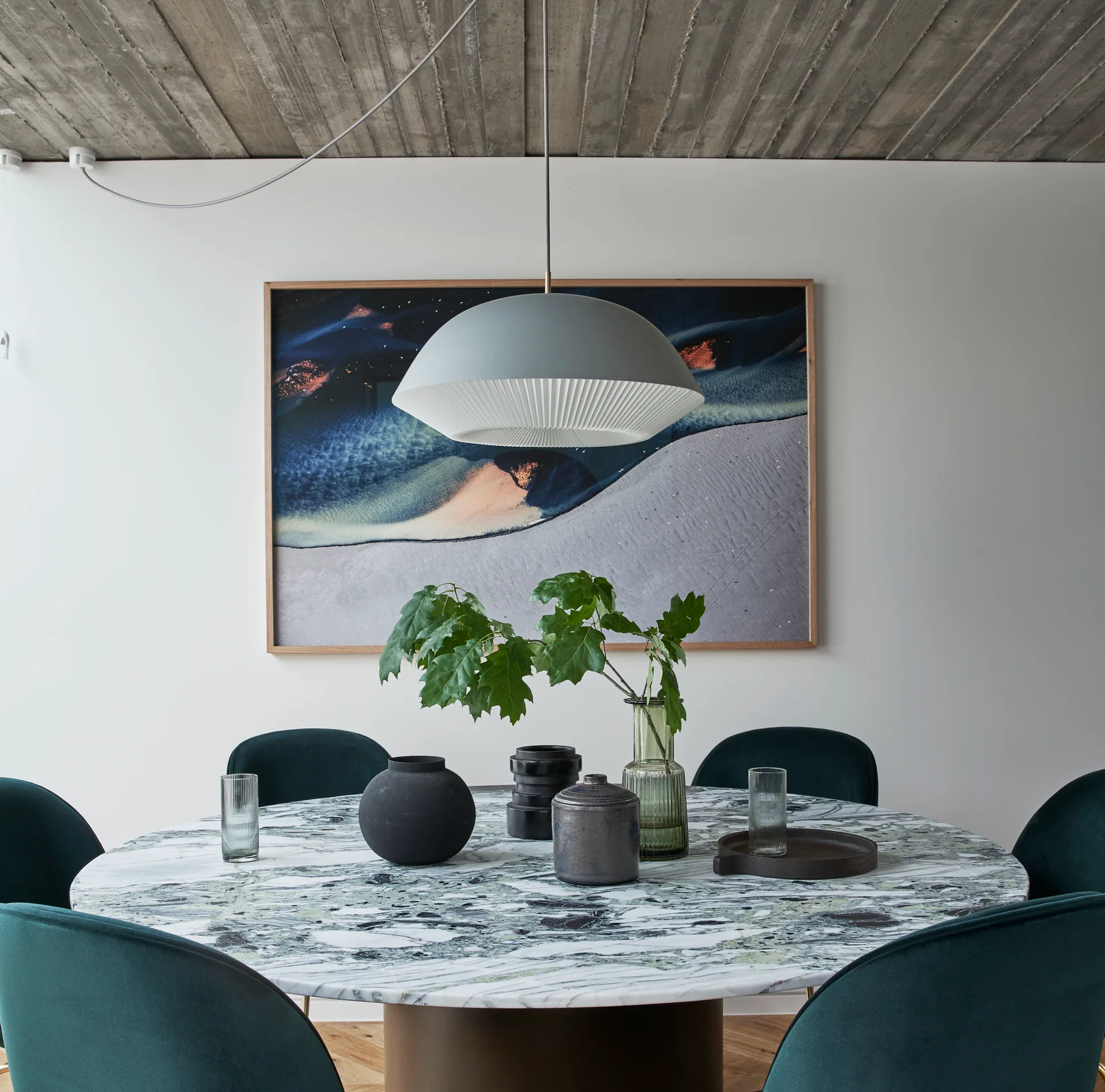 A round green marble dining table with velvet chairs sits beneath a grey dome pendant light and a ribbed concrete ceiling.