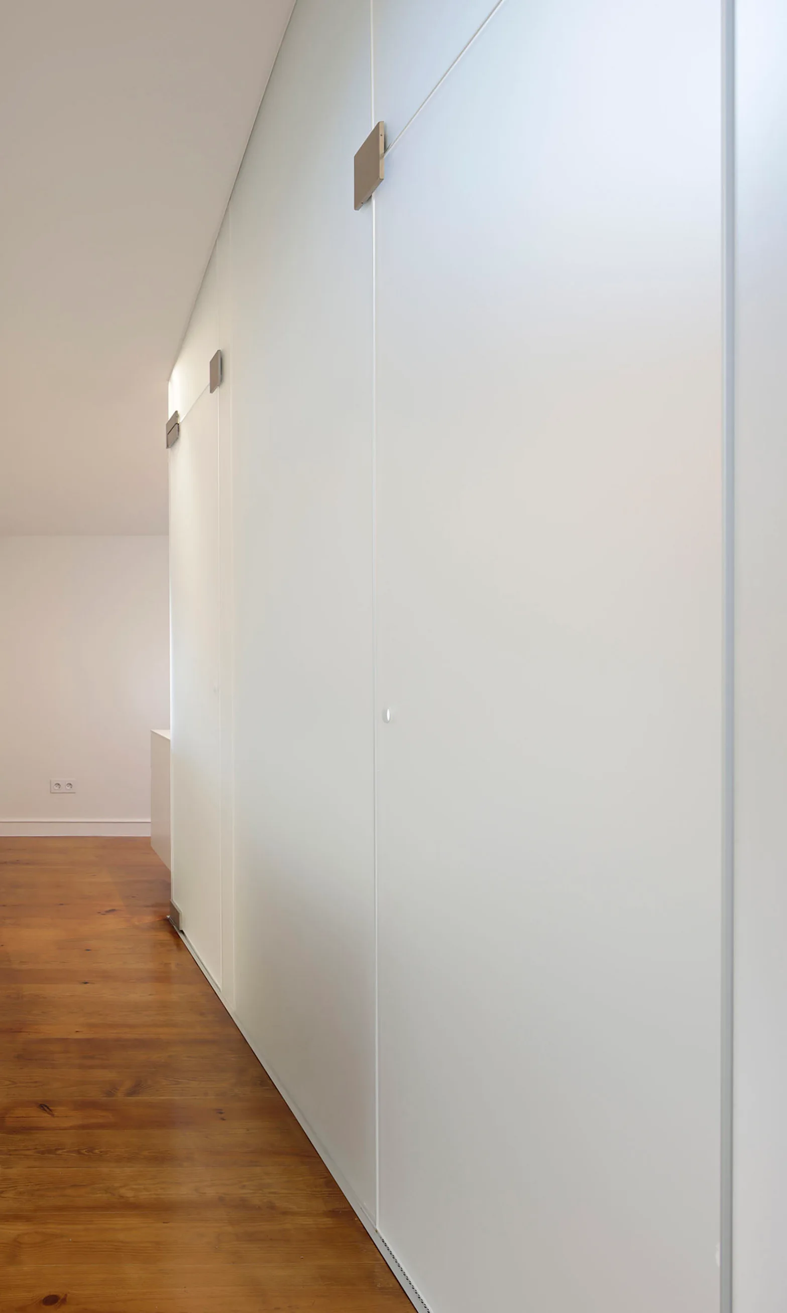 Minimalist white floor to ceiling storage cabinets line a hallway with warm brown wood plank flooring under a plain white ceiling.