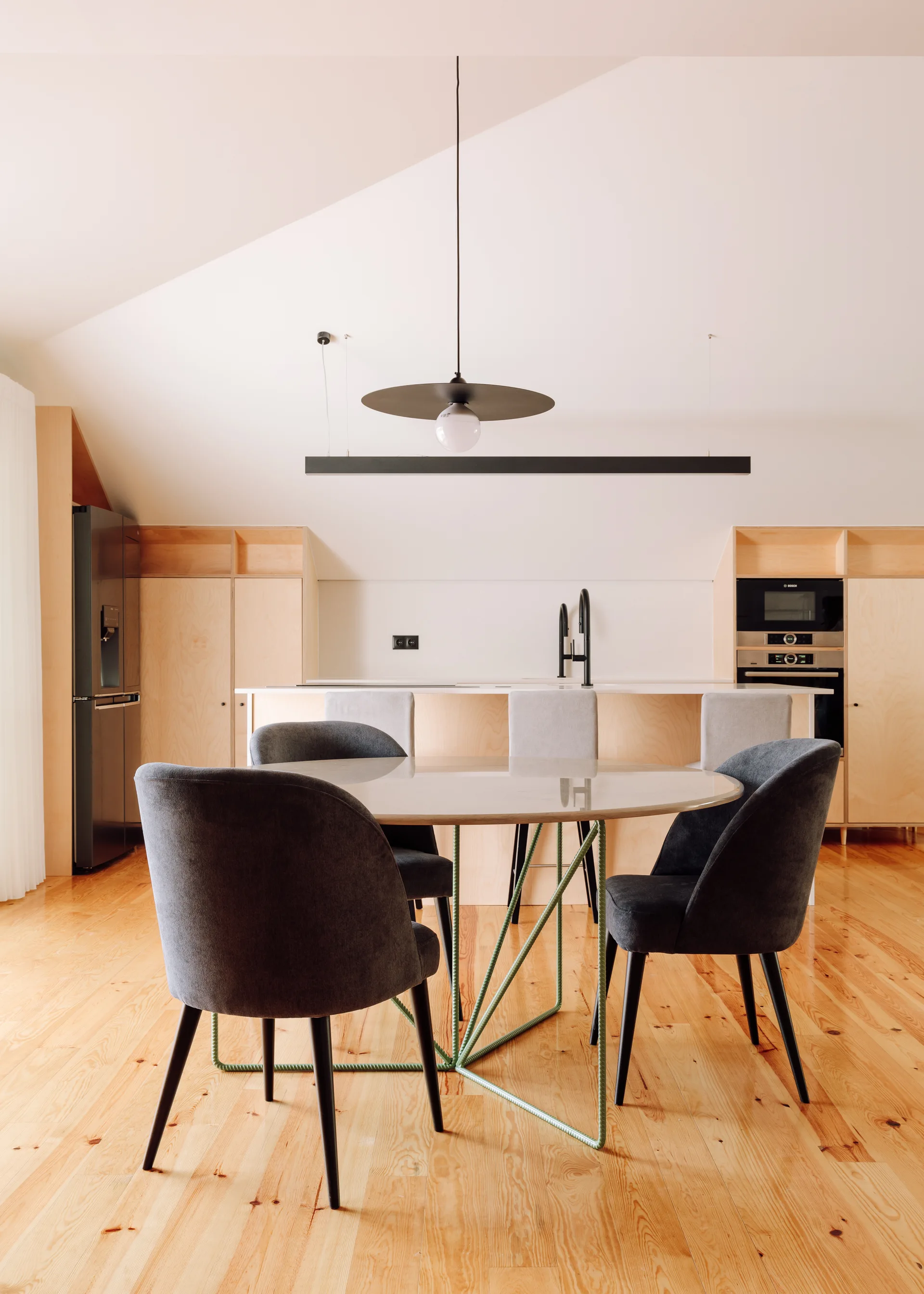A modern kitchen and dining area featuring light wood cabinetry, a round dining table with dark grey chairs, and a slanted white ceiling.