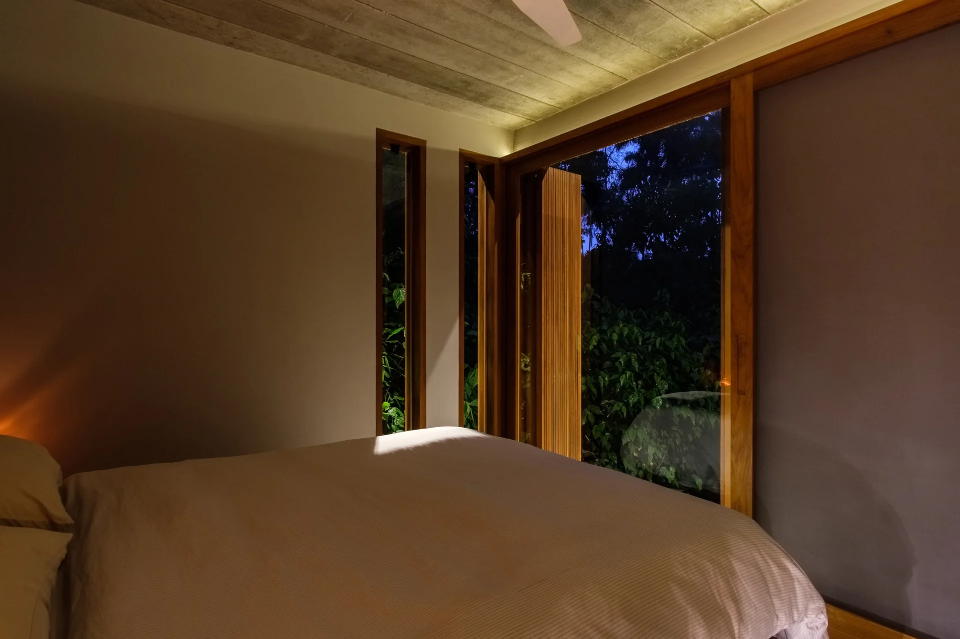 A bed with light bedding is positioned next to large wood-framed glass windows looking out to a dark garden under an exposed concrete ceiling.