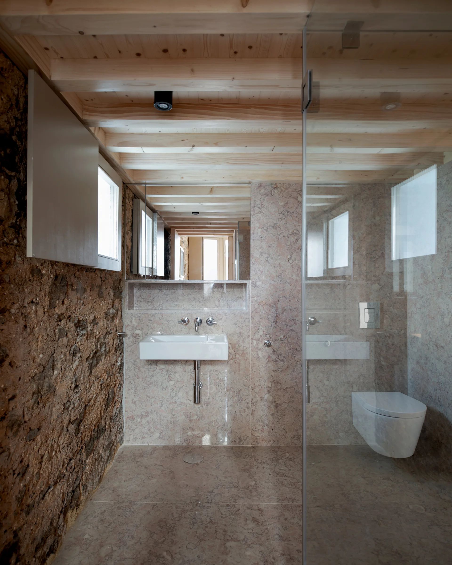 A modern bathroom featuring polished pink stone walls, a wall-mounted sink, a glass shower partition, and an exposed timber ceiling.