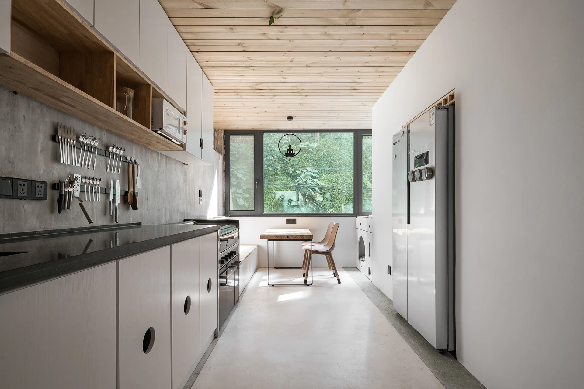 A modern galley kitchen featuring a natural wood ceiling, white cabinetry with circular cutouts, and a concrete wall leading to a window dining nook.