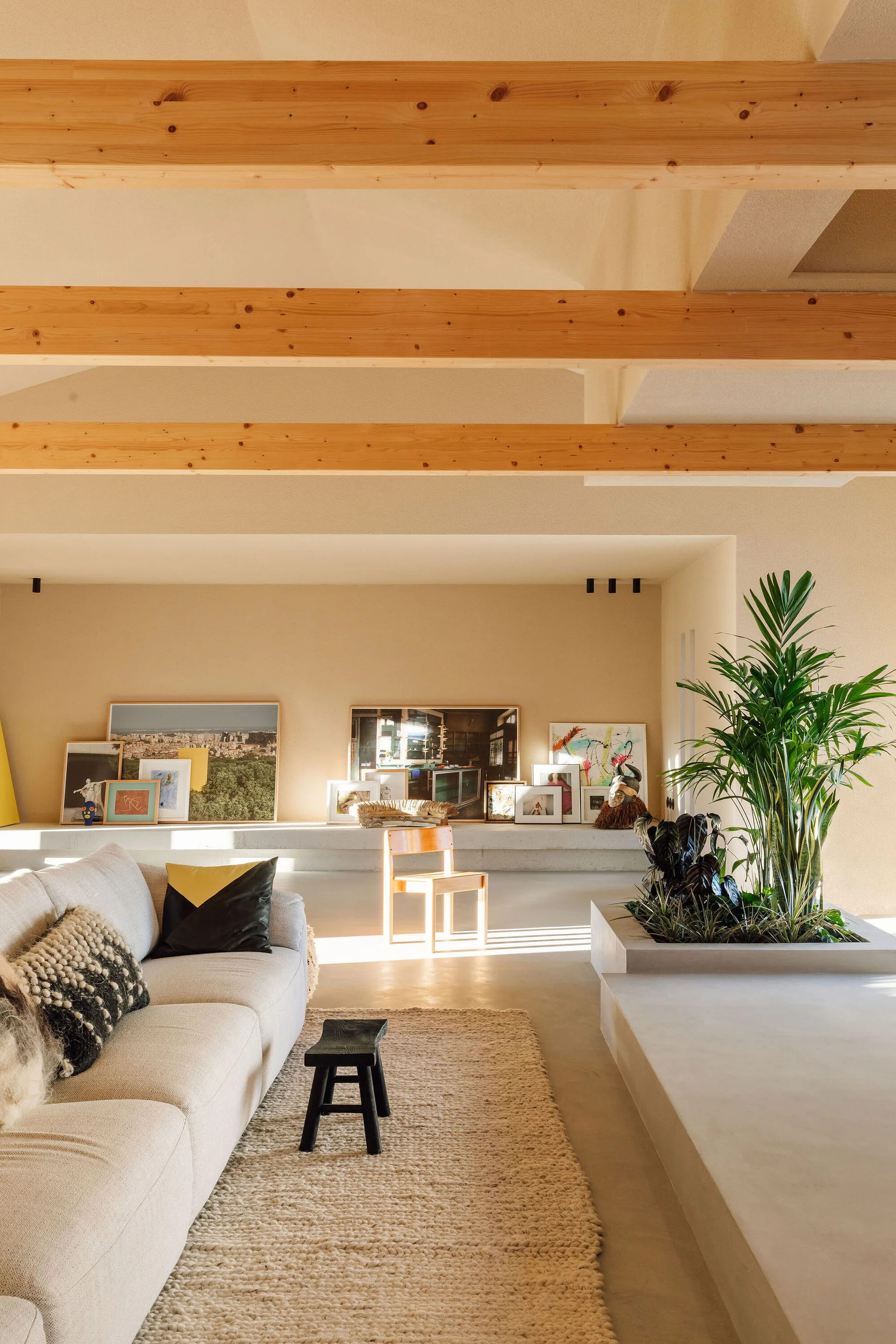 A minimalist living room features exposed wood ceiling beams, a cream sofa on a textured rug, and a low concrete ledge displaying leaning artwork.