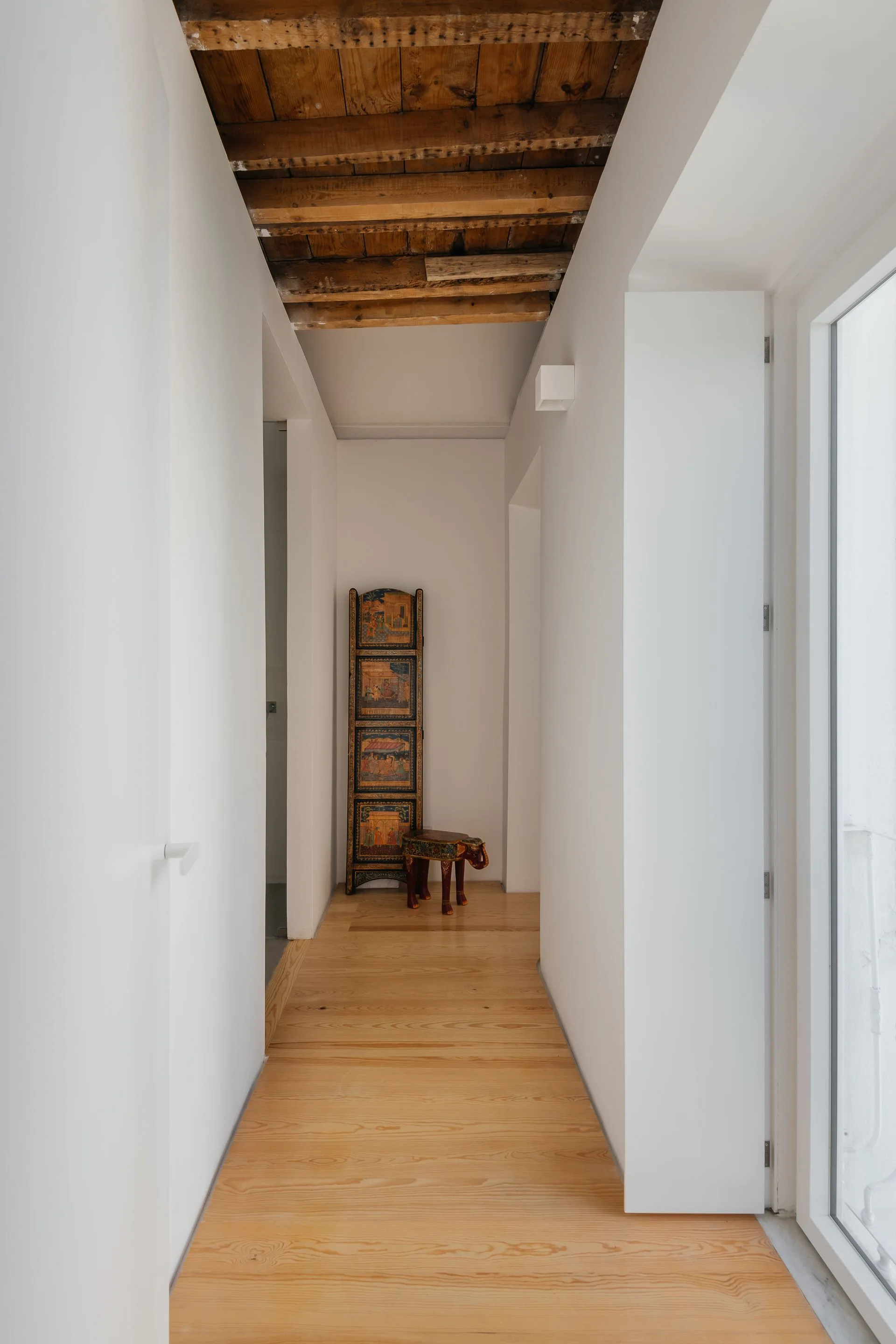A narrow white hallway with light wood floors features an rustic exposed wood beam ceiling and a decorative folding screen at the far end.