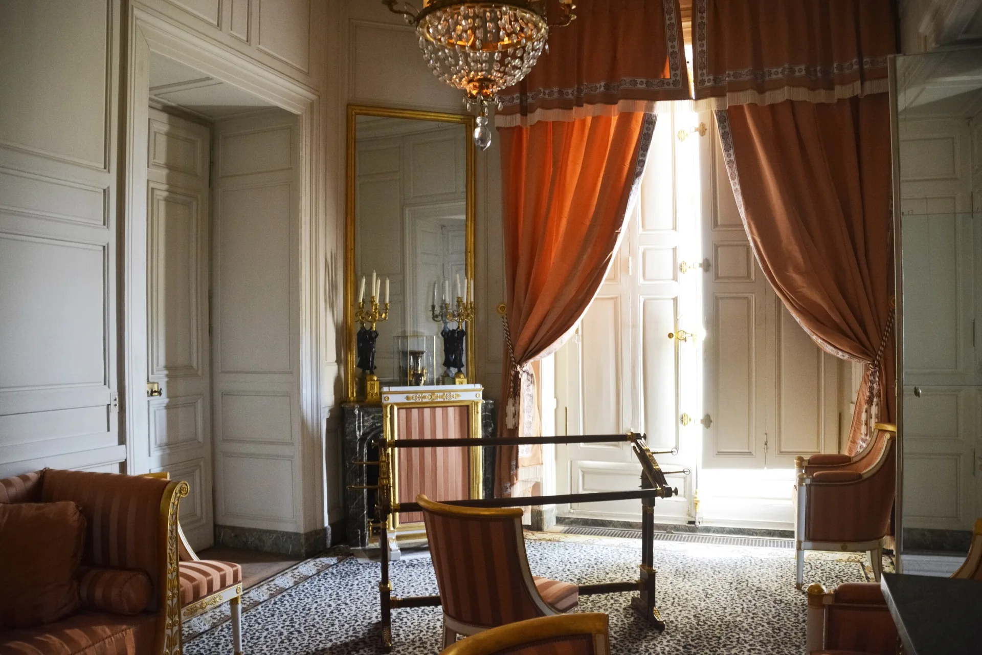 A historic parlor featuring peach curtains, striped armchairs, a marble fireplace, and a leopard print carpet under a crystal chandelier.