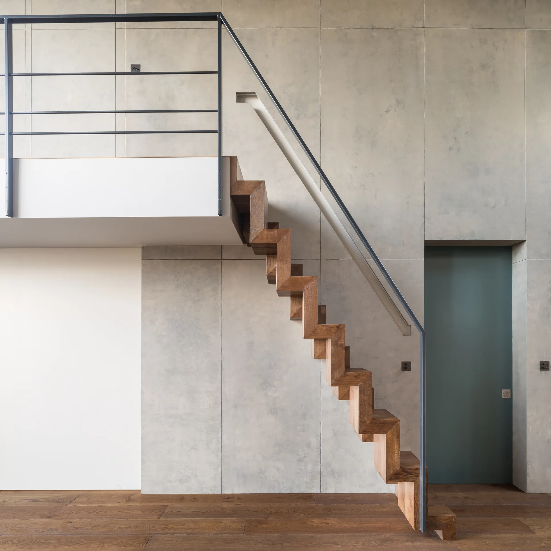 A geometric wooden zig-zag staircase with black metal handrails set against a grey concrete-paneled wall next to a teal door.