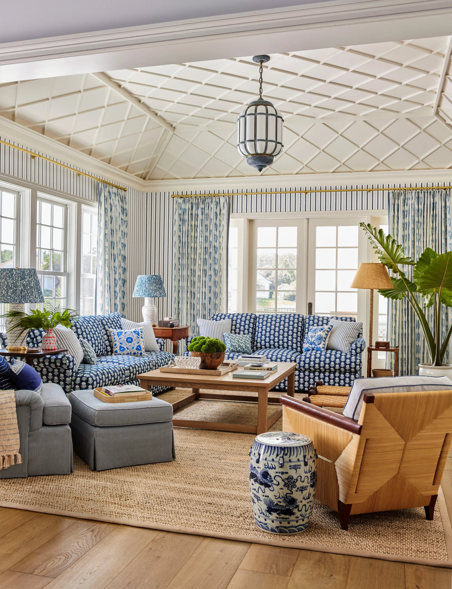 A bright family room featuring blue and white patterned sofas, striped wallpaper, a jute rug, and a geometric lattice ceiling.