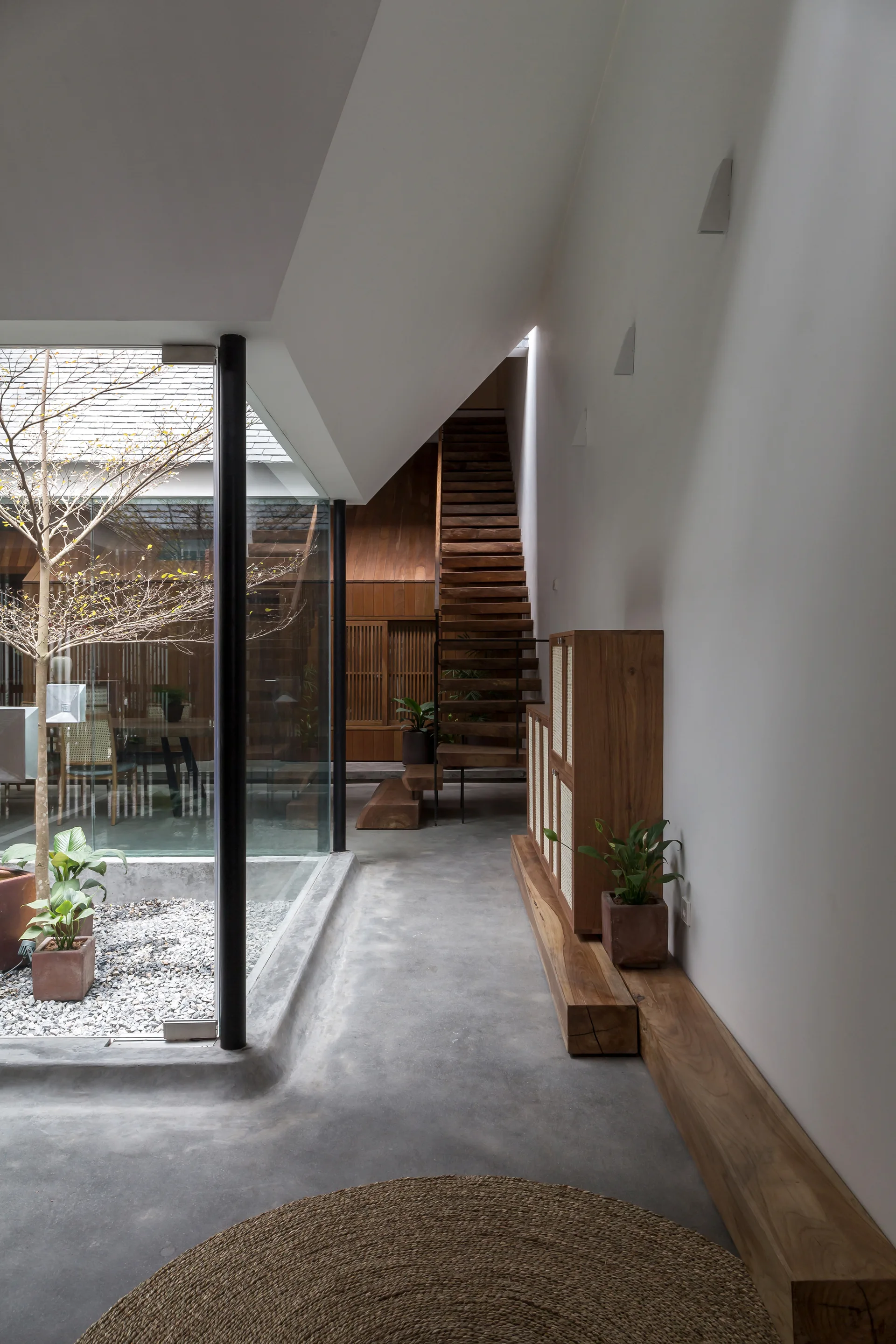 A polished concrete hallway features a glass-walled internal courtyard on the left and a wooden staircase and cane-front cabinet on the right.