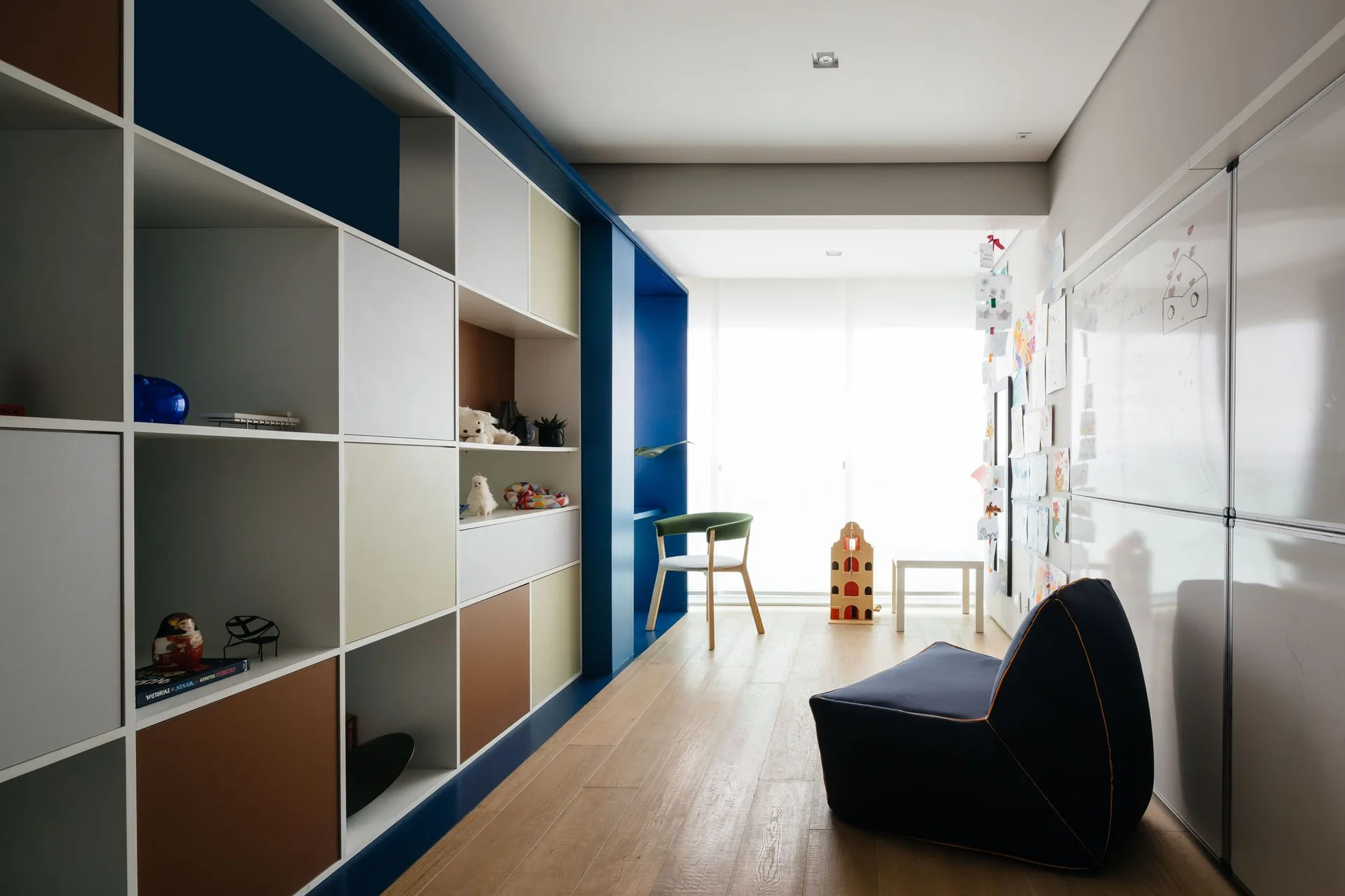 A modern kids playroom features a large color-blocked built-in shelving unit, a whiteboard wall, a blue bean bag, and light wood flooring.