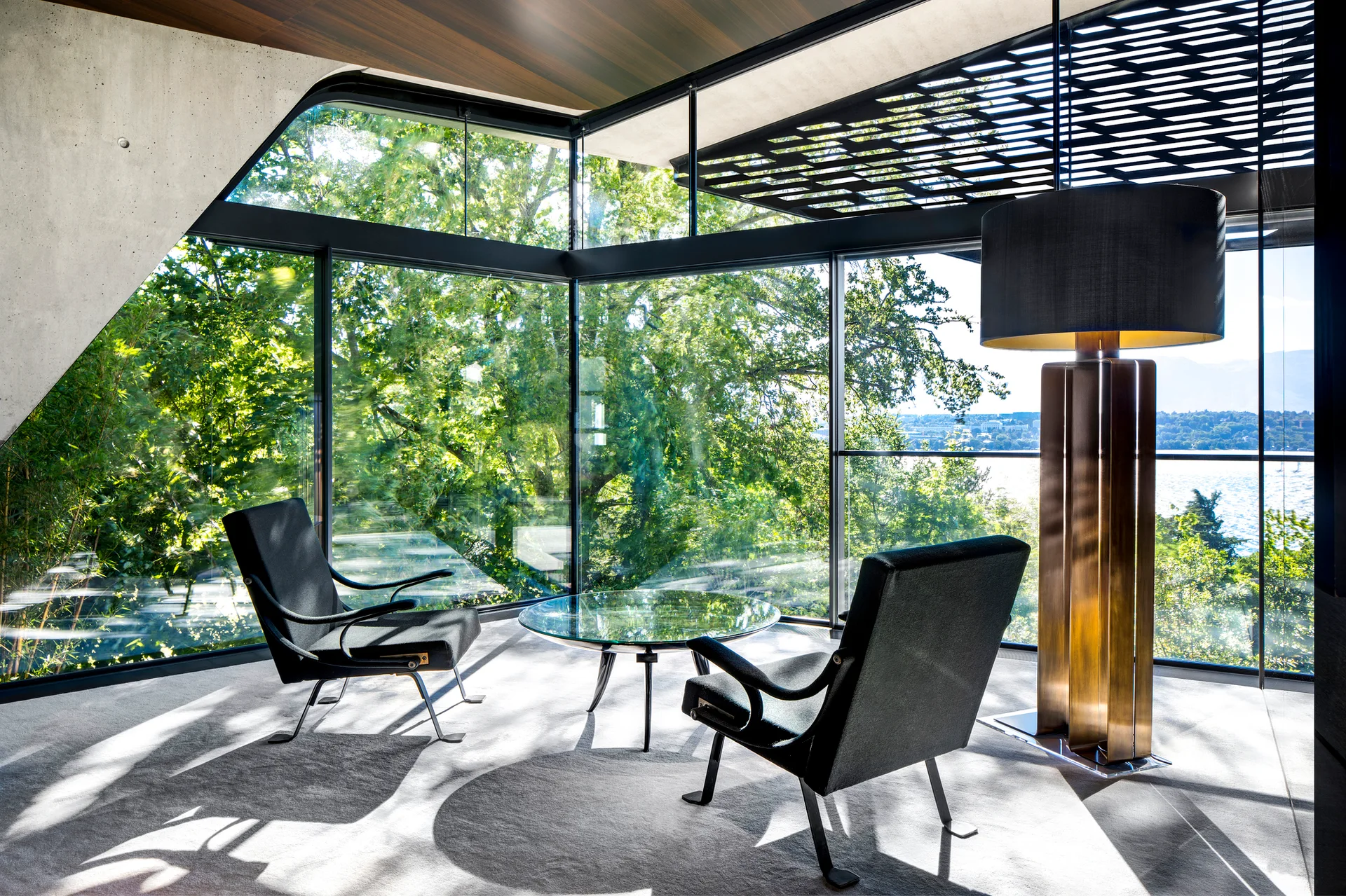 A modern minimalist lounge area features two black armchairs and a glass coffee table in front of floor-to-ceiling windows showing green trees.