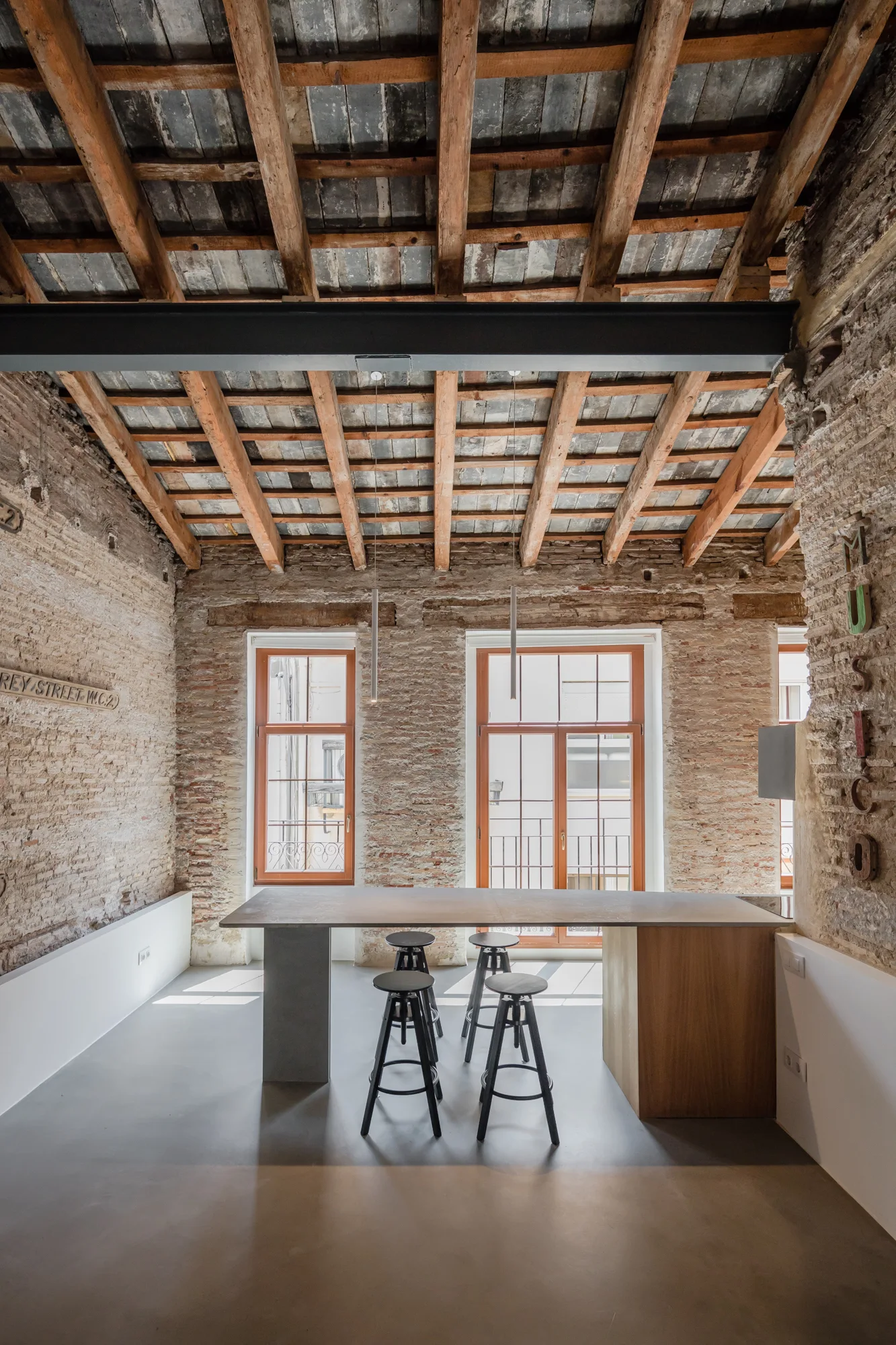 An industrial kitchen featuring exposed brick walls, a wooden beamed ceiling, and a long island with black stools.