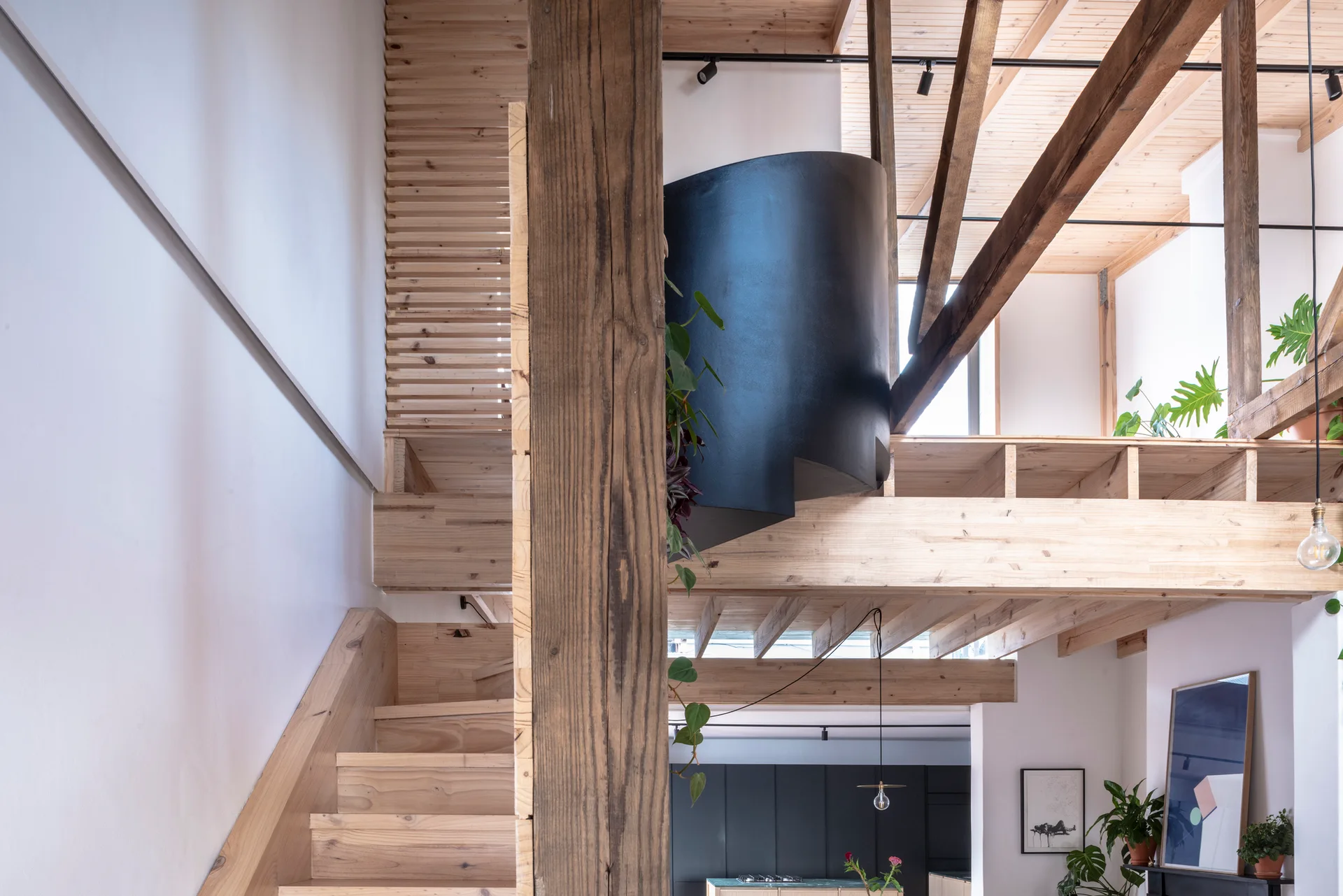 An open loft space features a light wood staircase, exposed ceiling beams, a mezzanine level, and a contrasting matte black spiral staircase element.