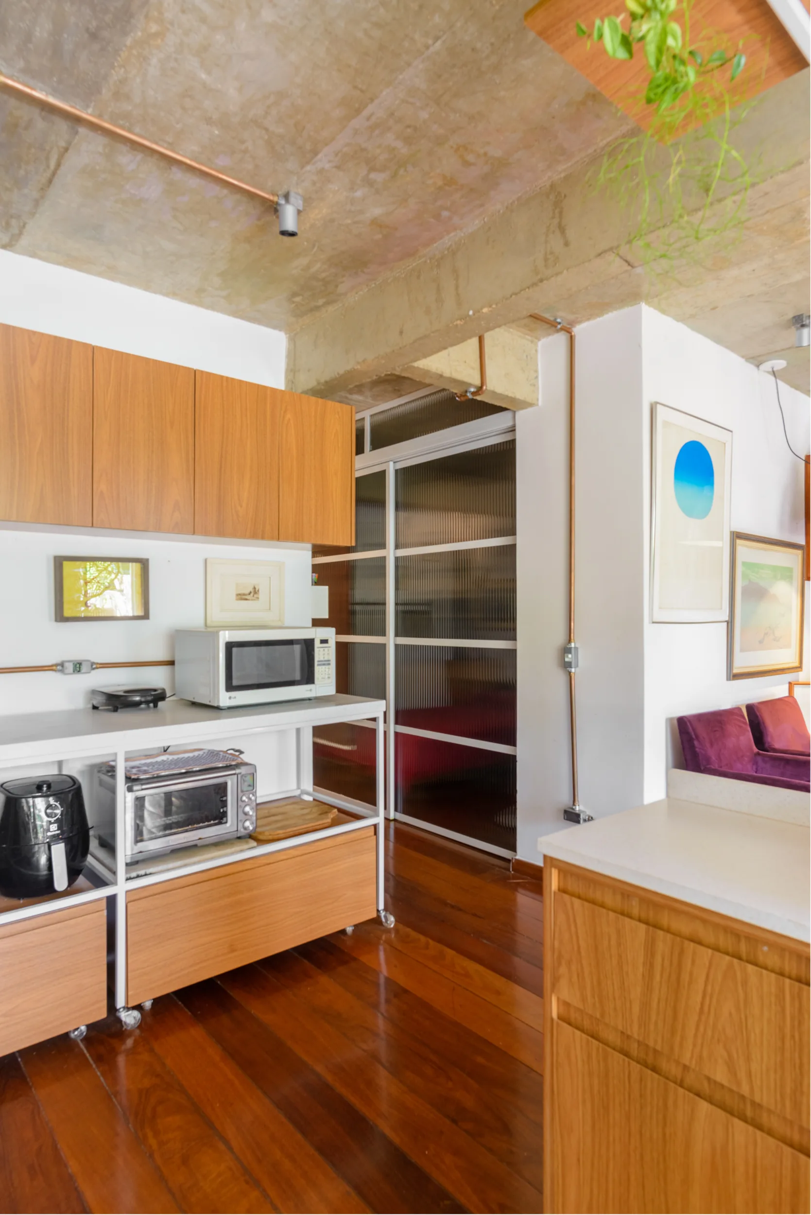 A modern industrial kitchen featuring wood cabinets, an exposed raw concrete ceiling with copper pipes, and a fluted glass sliding partition door.