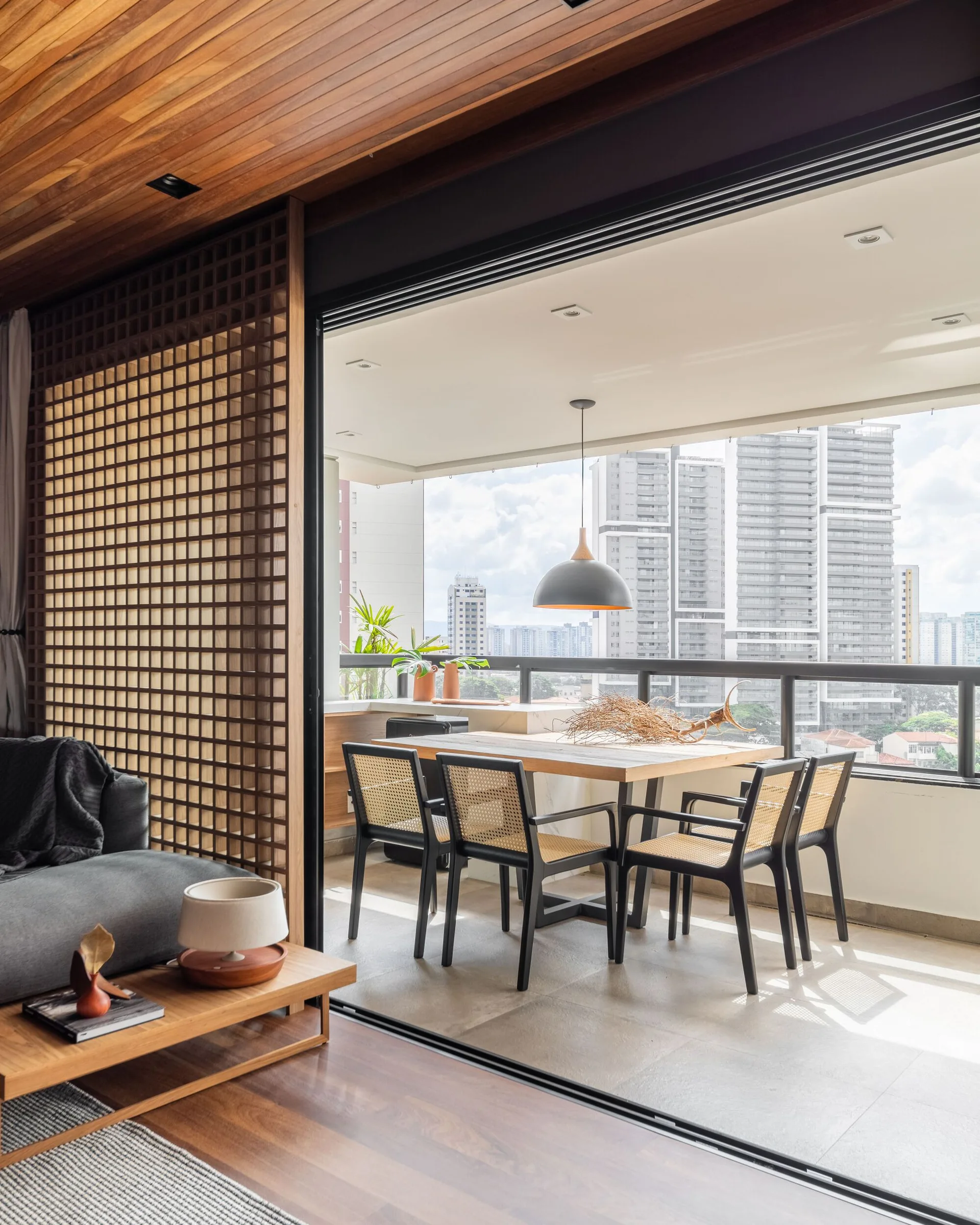 A modern balcony dining set with cane chairs overlooks a city skyline, connected to an indoor living space by sliding glass doors.