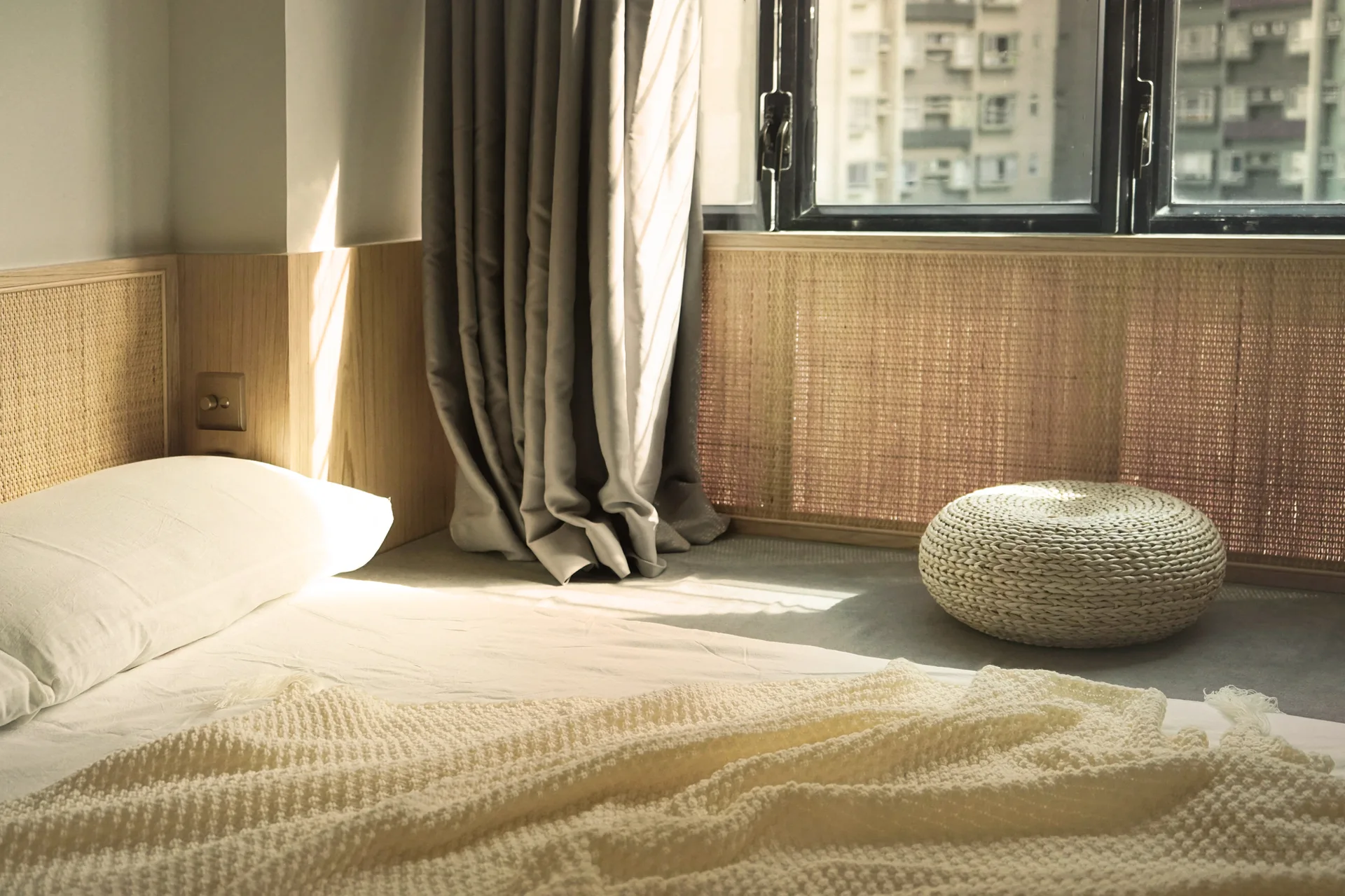 A sunlit bedroom with white bedding, a light wood and rattan headboard, and a matching rattan screen under a black-framed window.