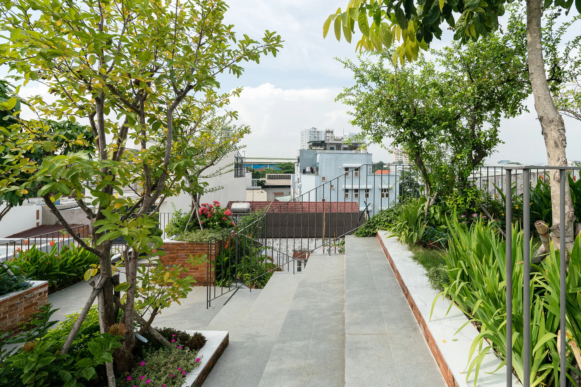 A rooftop terrace features wide grey stone steps alongside red brick planters filled with lush green trees and plants.