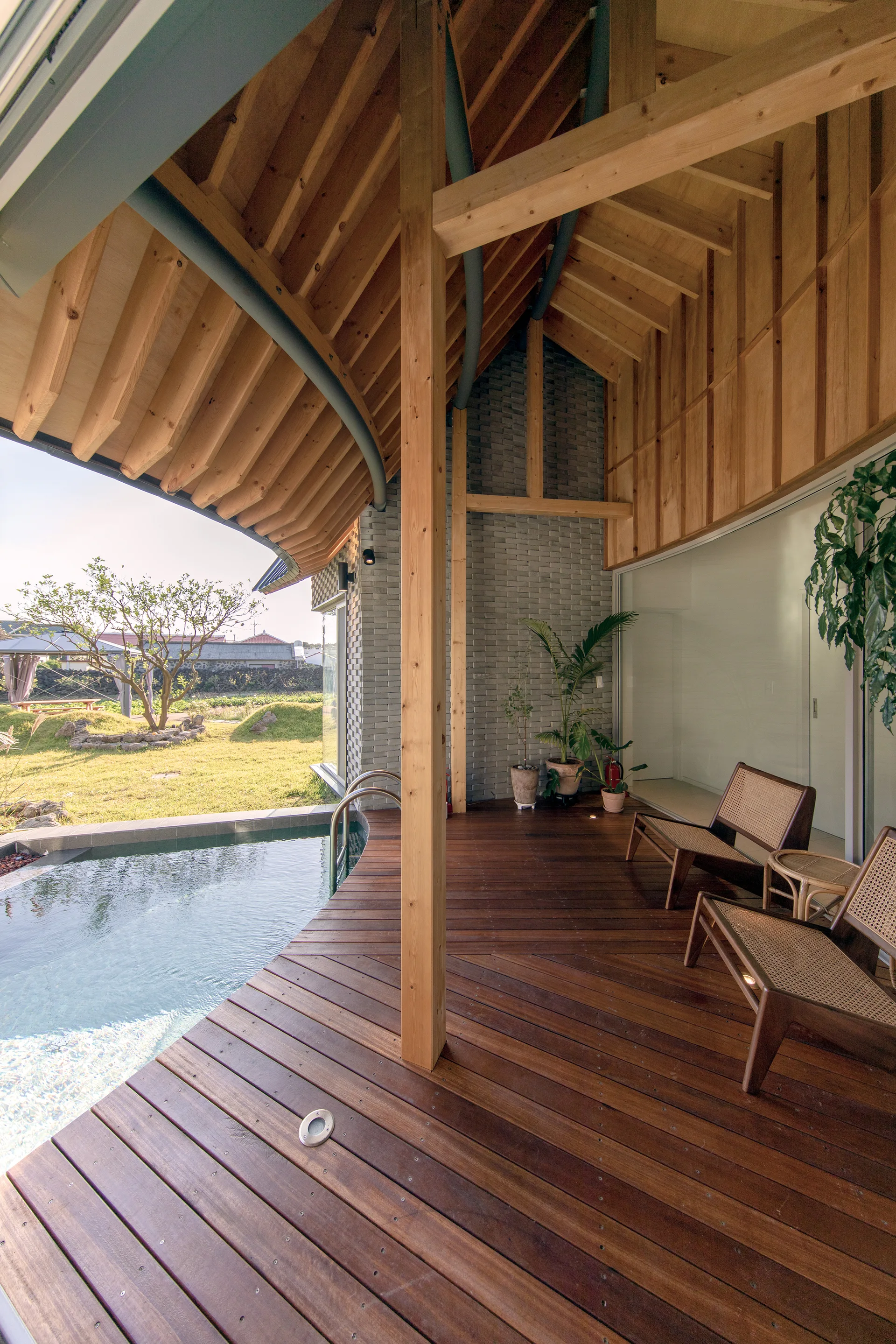 A covered wood deck featuring a curved swimming pool, exposed timber ceiling beams, a gray brick wall, and two wood and cane lounge chairs.