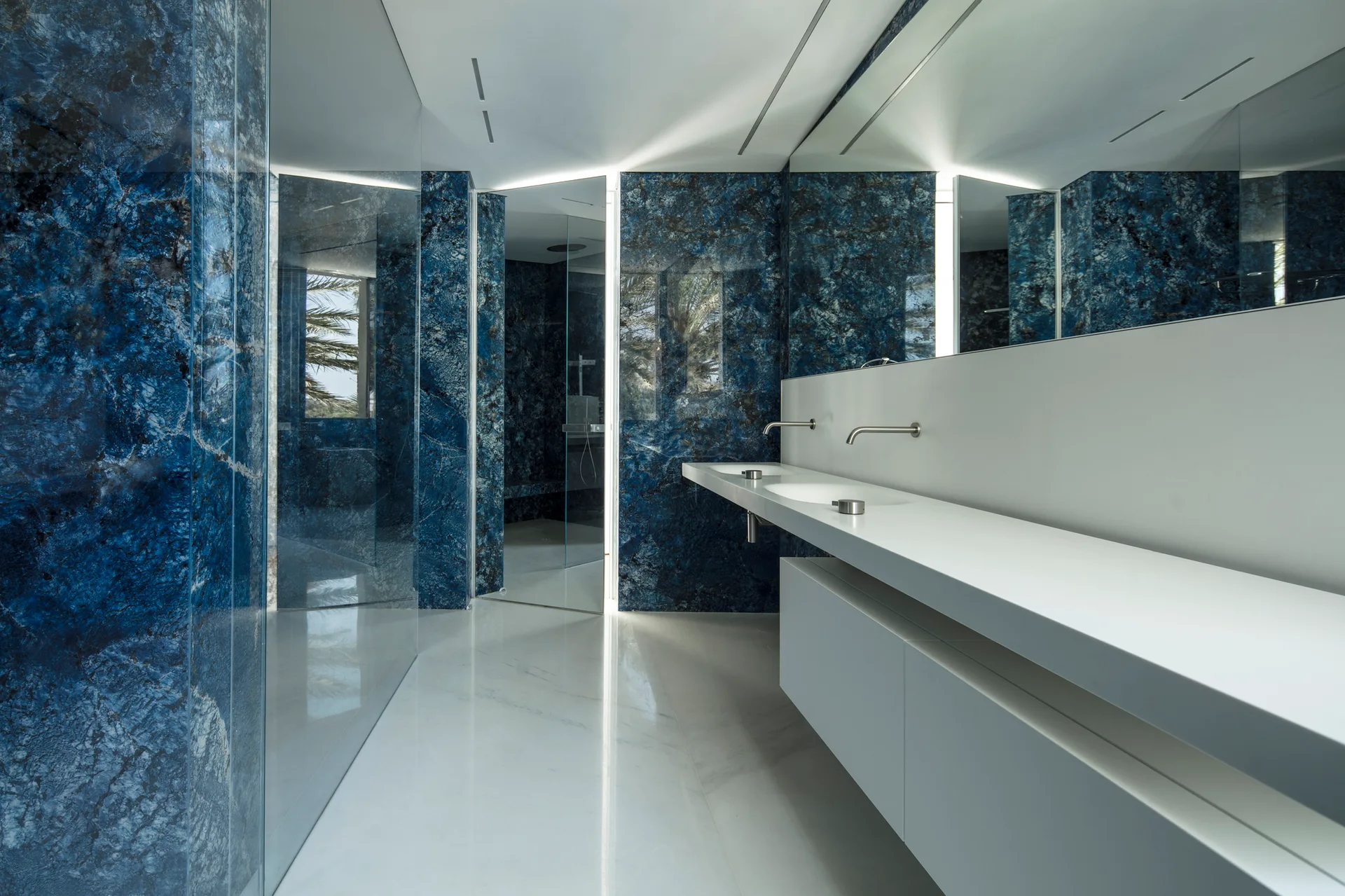 A modern bathroom featuring polished blue marble wall cladding, a floating white double vanity with wall-mounted faucets, and white floor tiles.