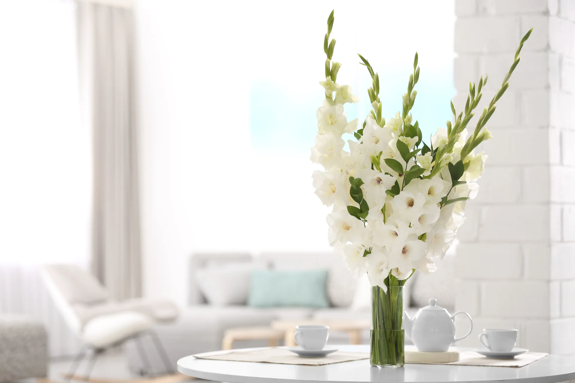 Tall white gladiolus flowers in a clear glass vase sit beside a white ceramic tea set on a round table, with a blurred living room in the background.
