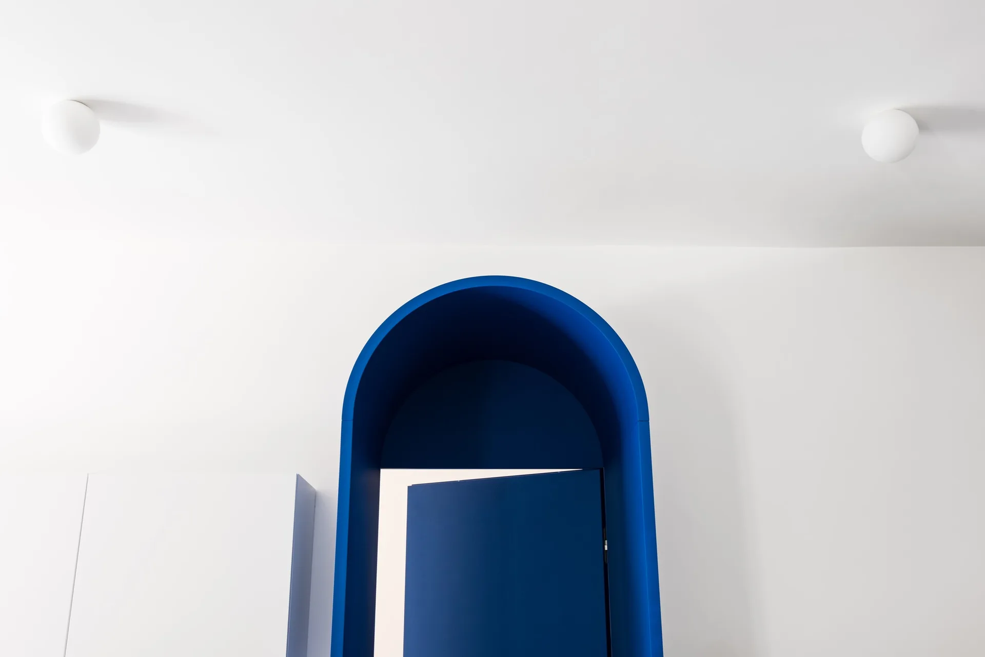 A minimalist interior features a stark blue arched doorway and door contrasted against white walls and ceiling with two globe lights.