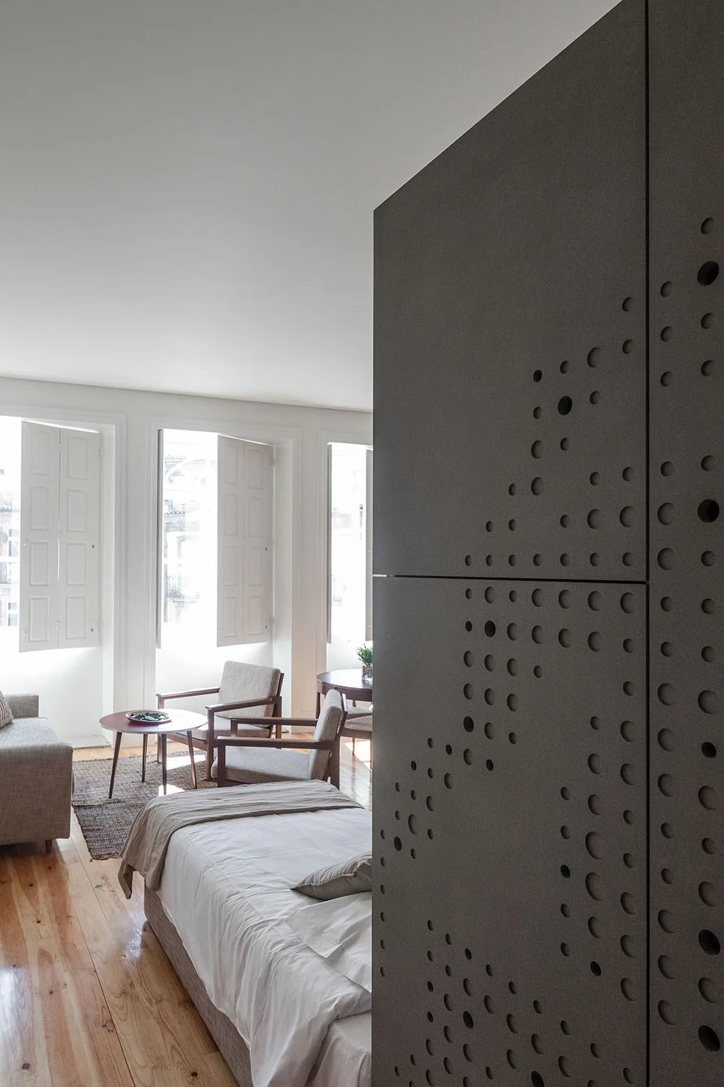 A contemporary bedroom featuring a large dark gray perforated room divider next to a bed and a sitting area with light wood floors.