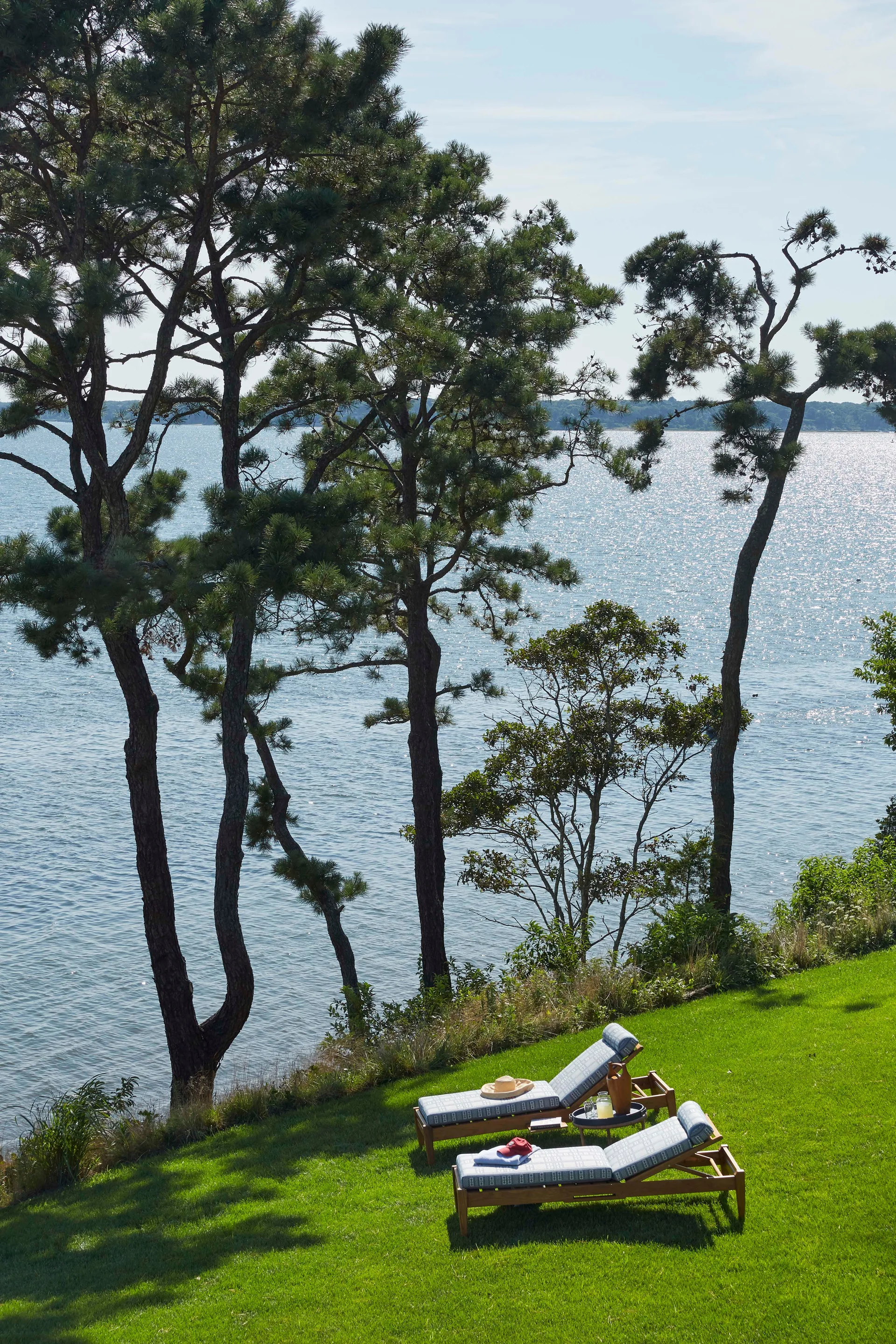 Two wooden chaise lounge chairs with patterned cushions sit on a green grassy lawn overlooking a calm body of water framed by tall pine trees.