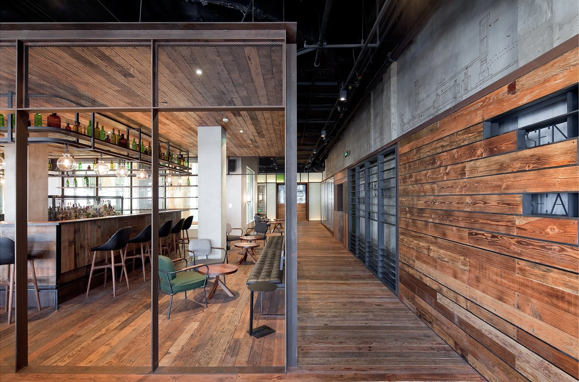 An industrial bar interior features weathered wood paneling on the walls and ceiling, a dark metal partition frame, bar stools, and a black open ceiling.