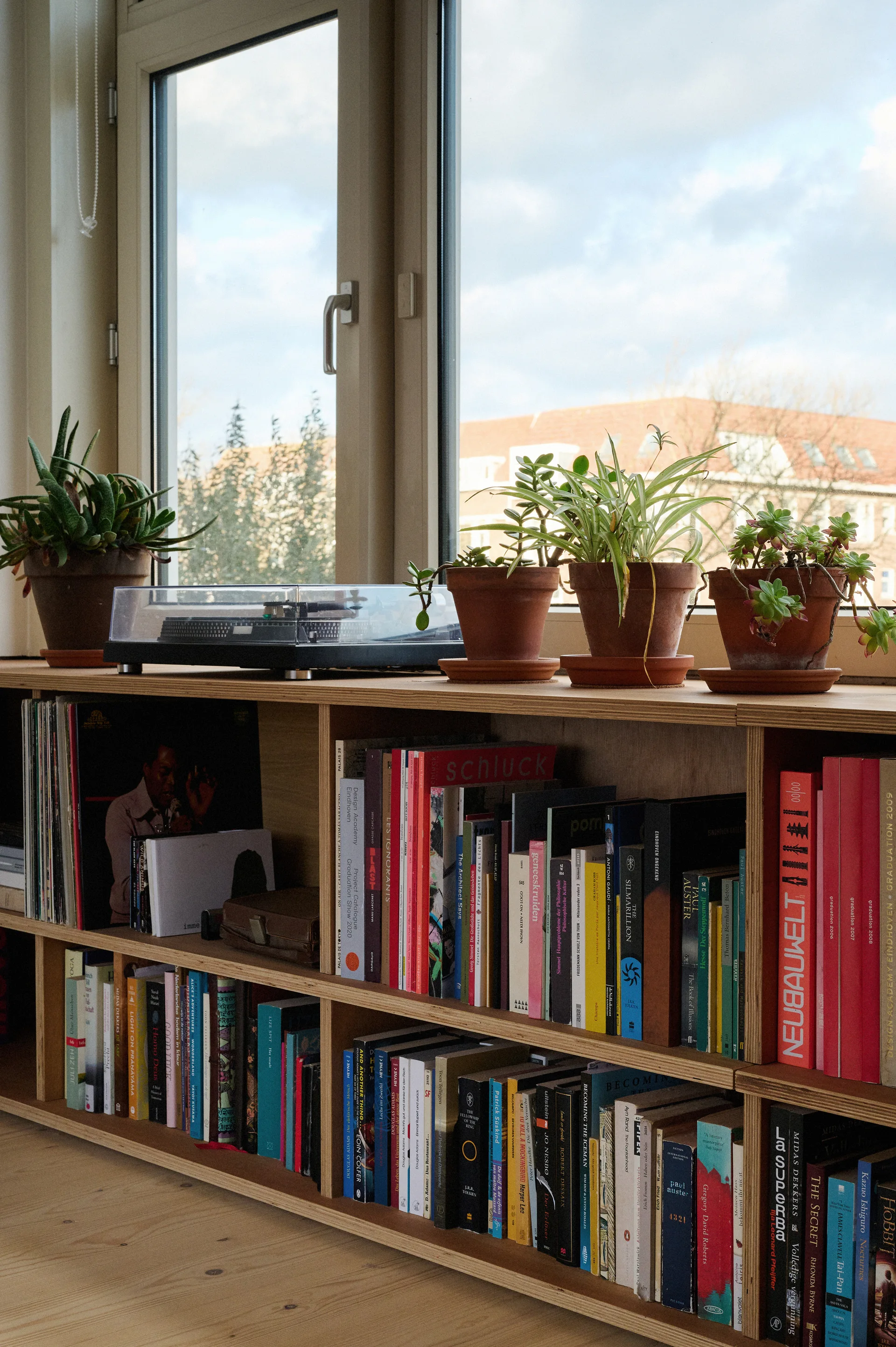 A low plywood bookshelf filled with books sits in front of a window, topped with a turntable and four potted plants in terracotta pots.