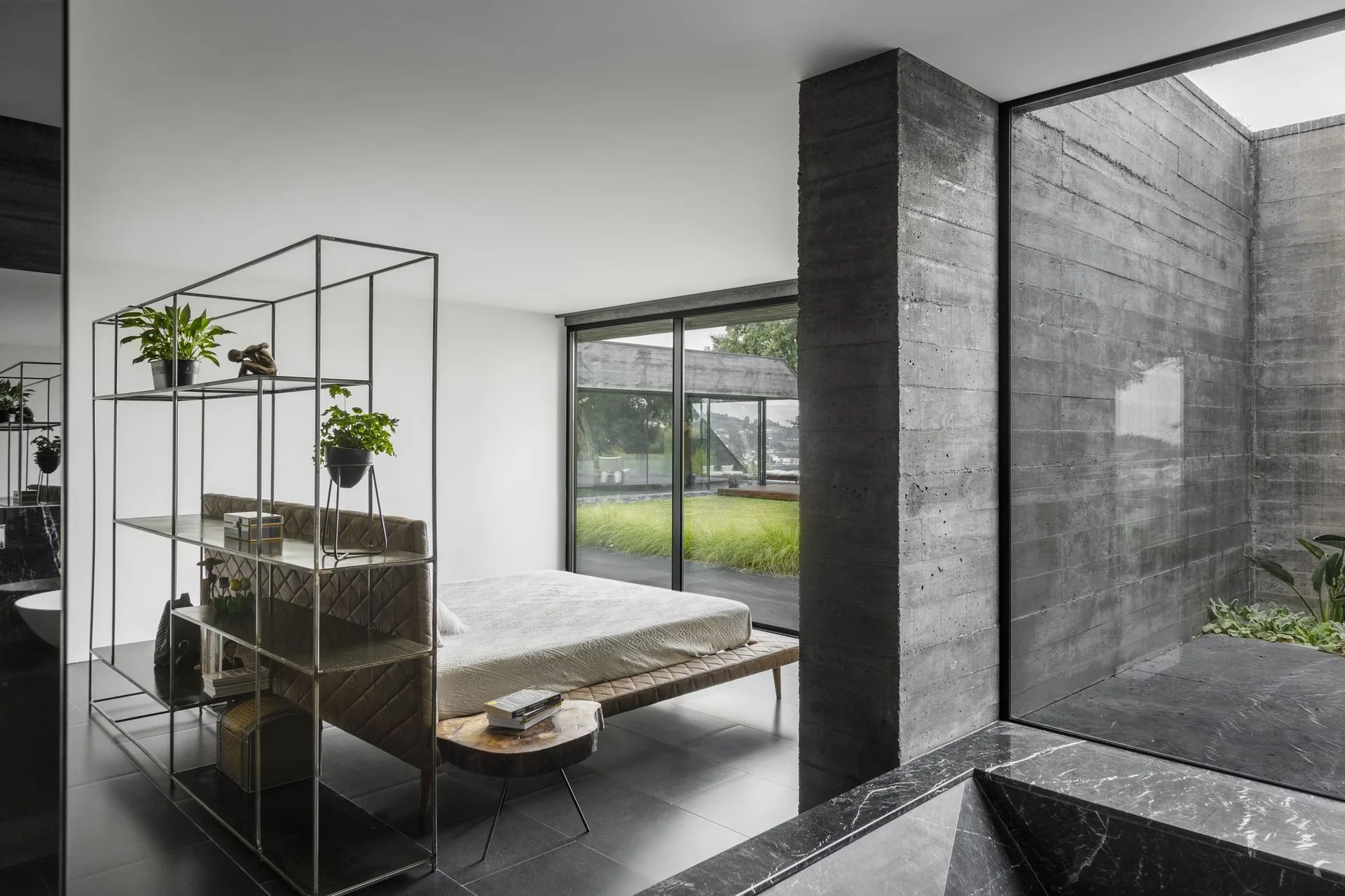A modern minimalist bedroom featuring a low quilted bed separated from a hallway by an open metal shelving unit next to floor-to-ceiling glass.
