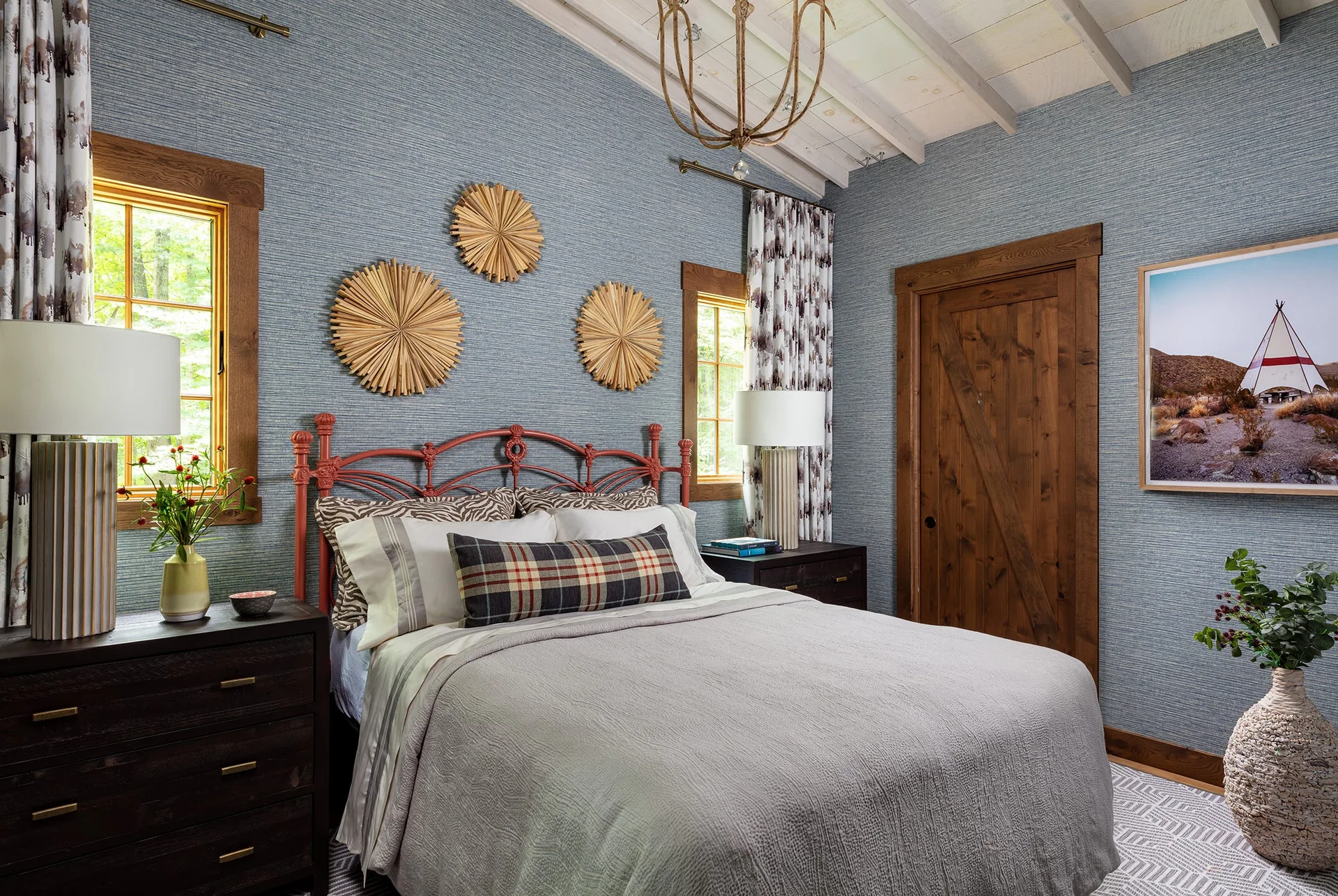 A rustic bedroom features blue grasscloth wallpaper, a terracotta-colored metal bed, dark wood nightstands, and a sliding wooden barn door.