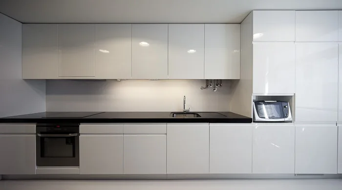 A minimalist kitchen featuring glossy white handleless cabinets, a black countertop with an integrated sink and oven, and a built-in microwave nook.