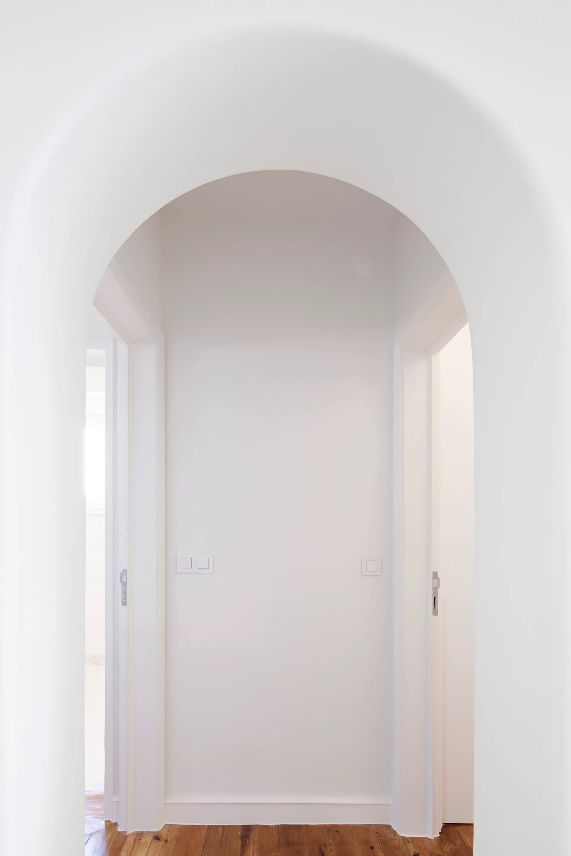 A view through a smooth white plaster archway into a minimalist hallway with natural wood plank flooring and white doorways.