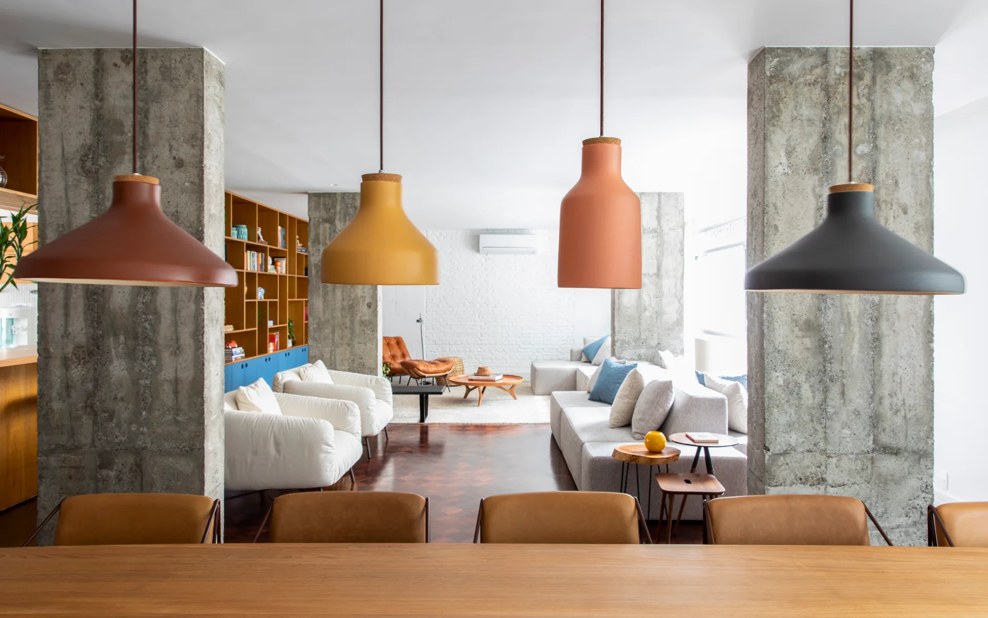 An open-plan living and dining room featuring exposed concrete columns, colorful pendant lights, parquet flooring, and a grey sectional sofa.